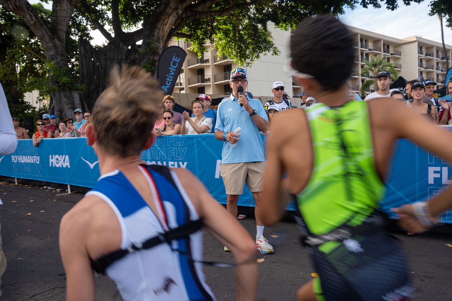 Marathon runners pass by spectators and volunteers at a race checkpoint with blue barriers along an urban course.