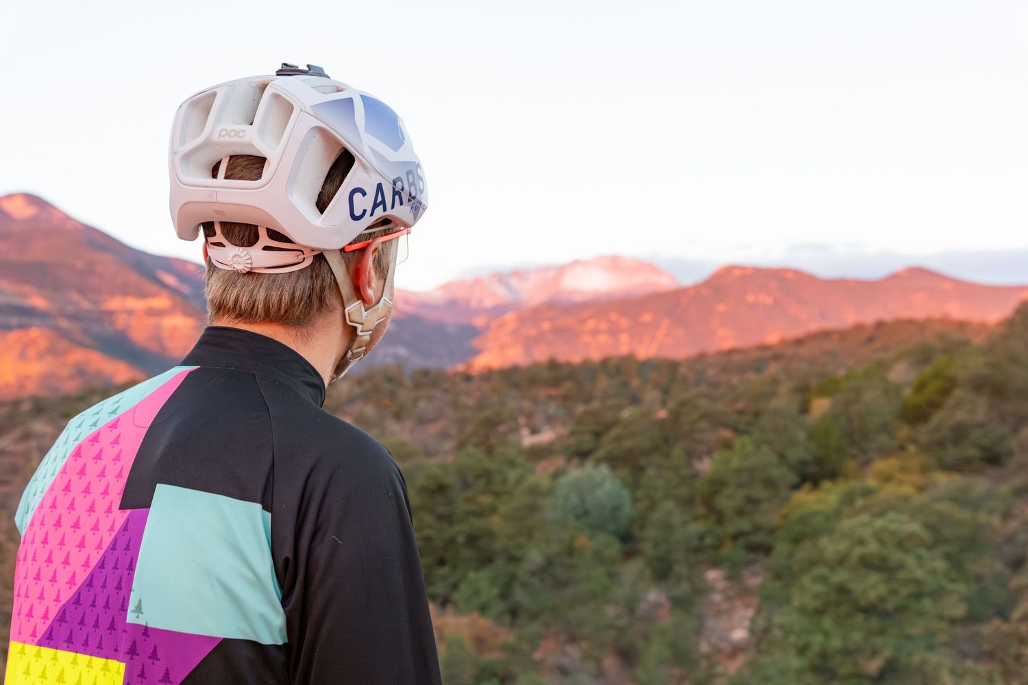 Rock climber in white helmet ascends steep cliff face against backdrop of red desert mountains at sunset.