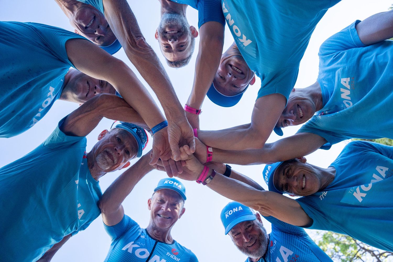 Group of people wearing light blue shirts huddle together in a circle formation looking down at the camera.