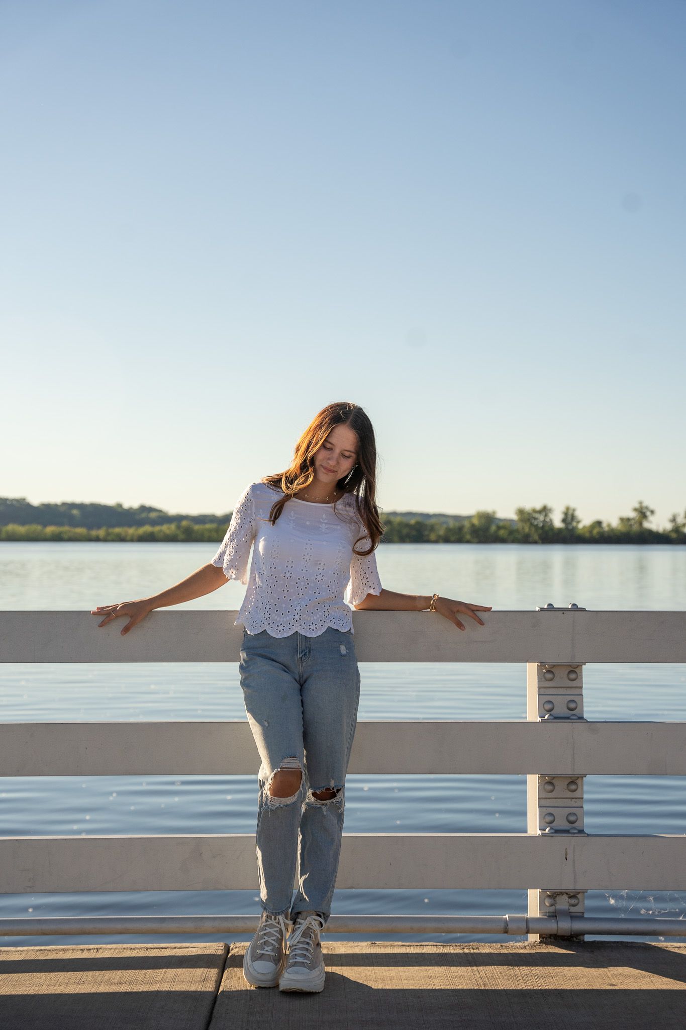 Person in ripped jeans and white top poses casually against a white railing by a serene lake at sunset.