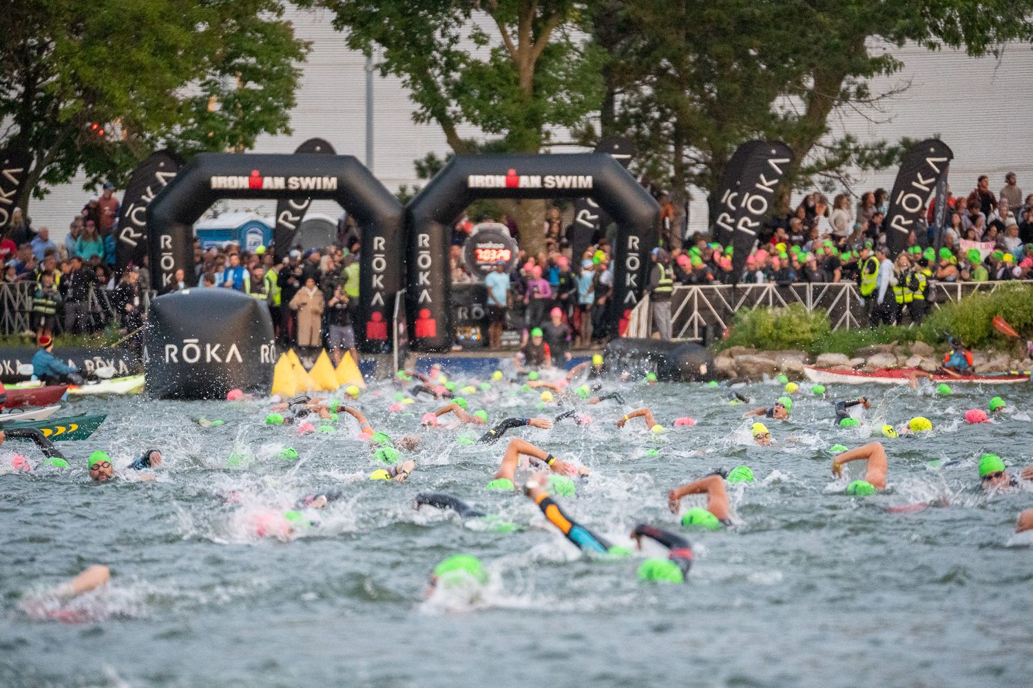 Swimmers compete in a triathlon race with colorful swim caps visible in the water near a crowded shoreline.