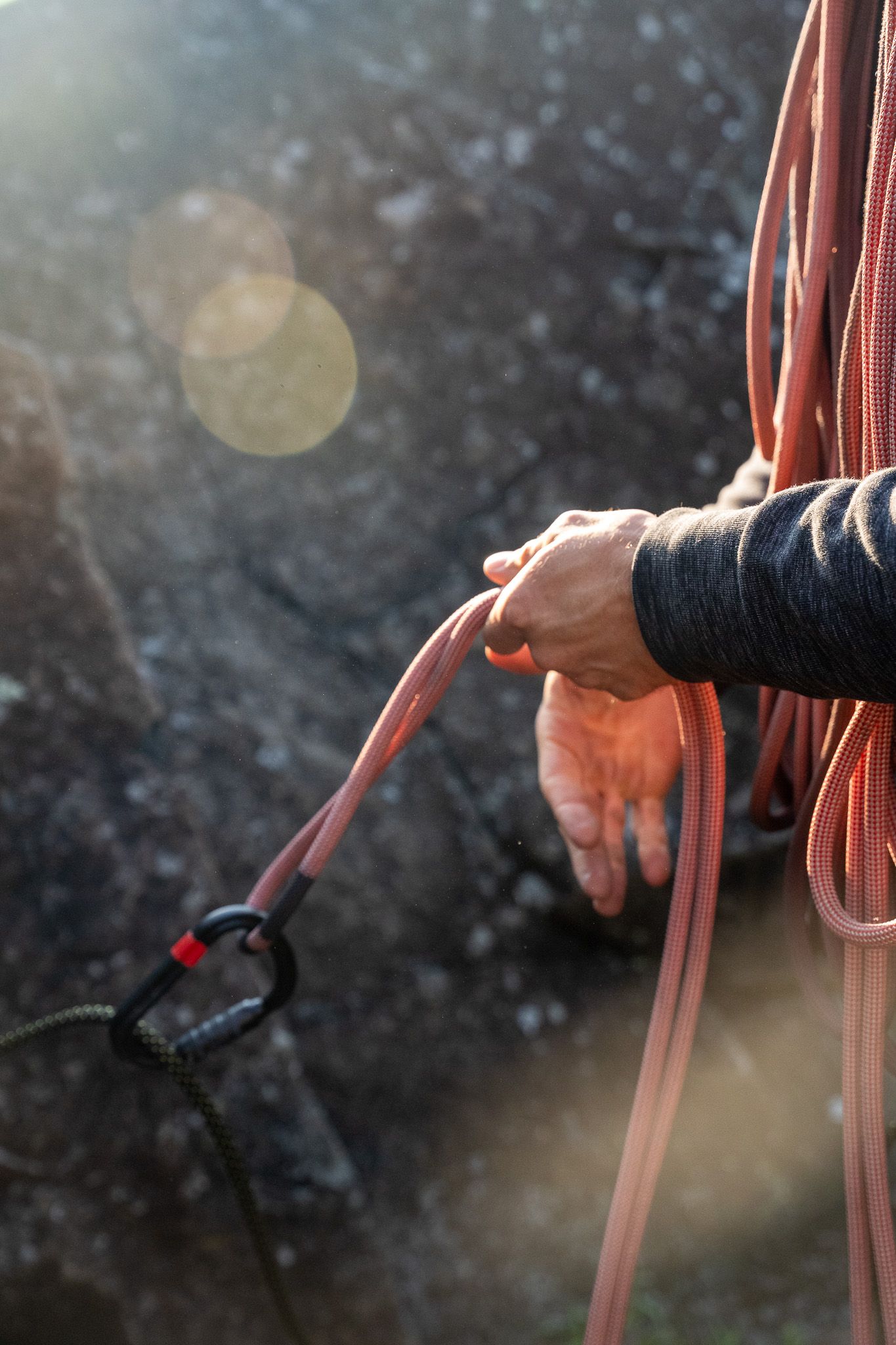 Close-up of a climber's hands holding rope and belay device with sunlit bokeh effect in background.