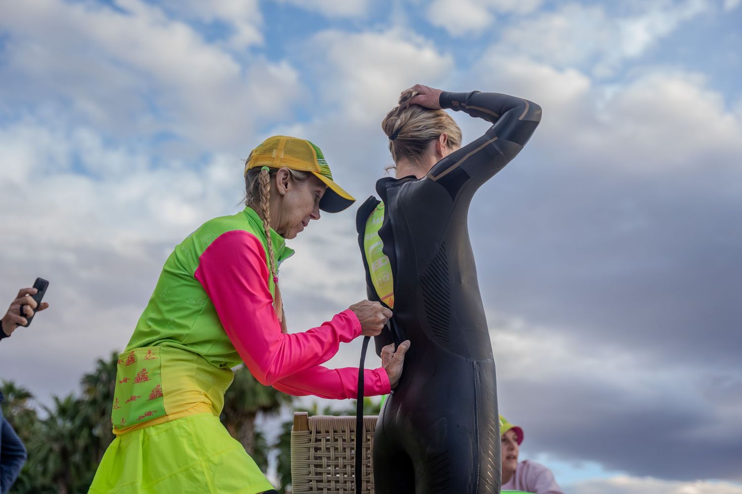 A triathlon participant gets help from a volunteer in a neon safety vest to put on their wetsuit before a race.