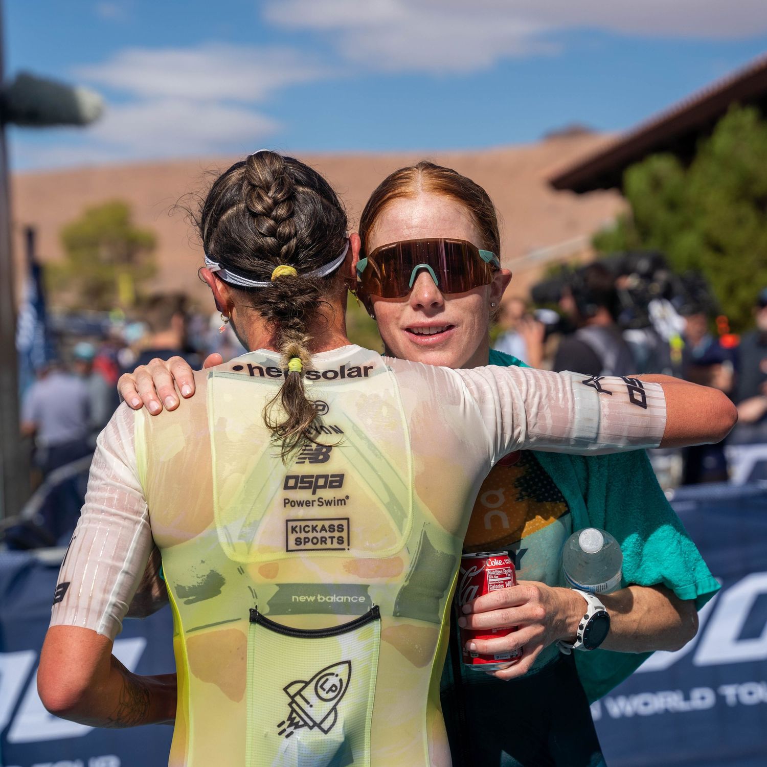 Two cyclists embrace after a race in front of desert hills with spectators gathered around them in the background.