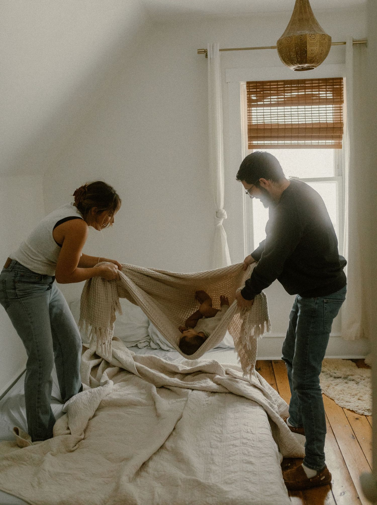 Parents playfully swinging their child in a white blanket hammock in a sunlit room with wooden floors.