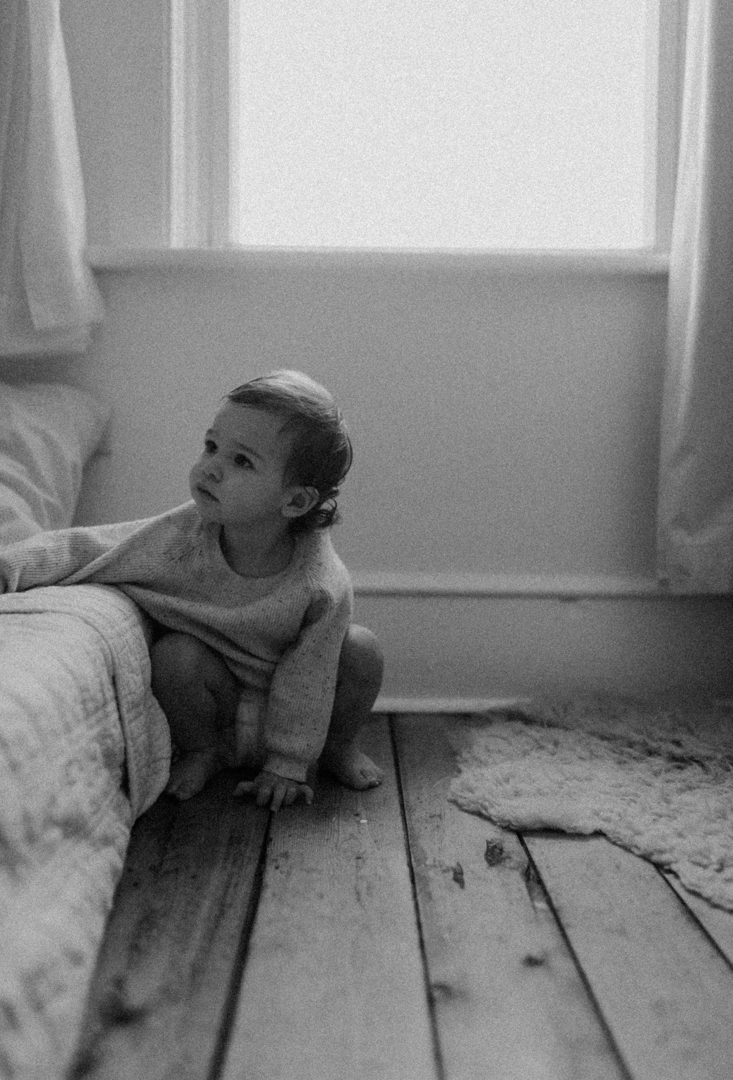 A small child explores beside a bed on rustic wooden flooring in black and white.