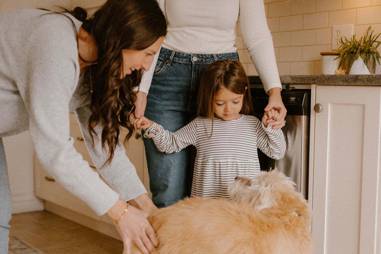 Two people and a small child pet a fluffy golden dog in a bright kitchen.