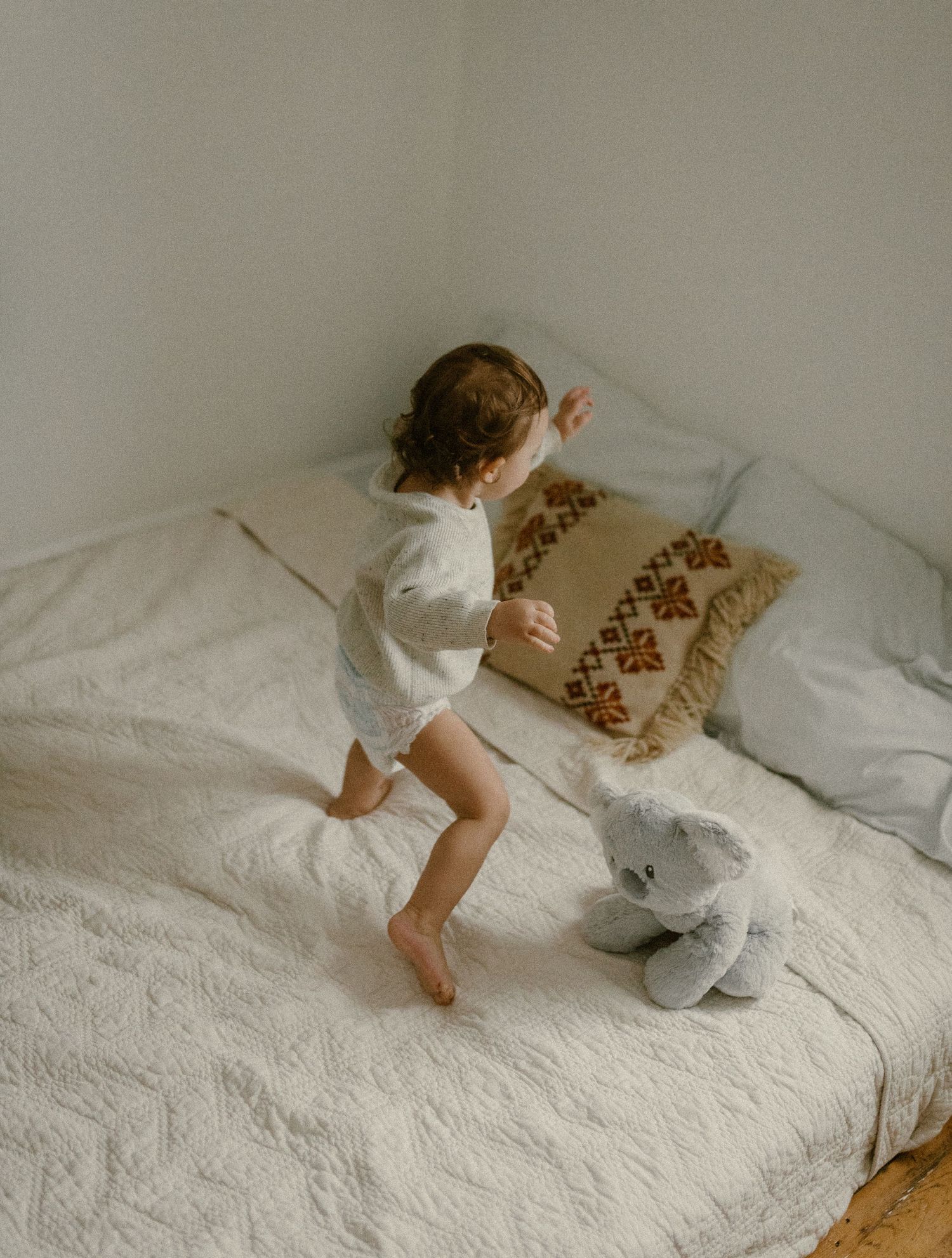 Small child playing on a white bed with decorative pillows and stuffed animal.