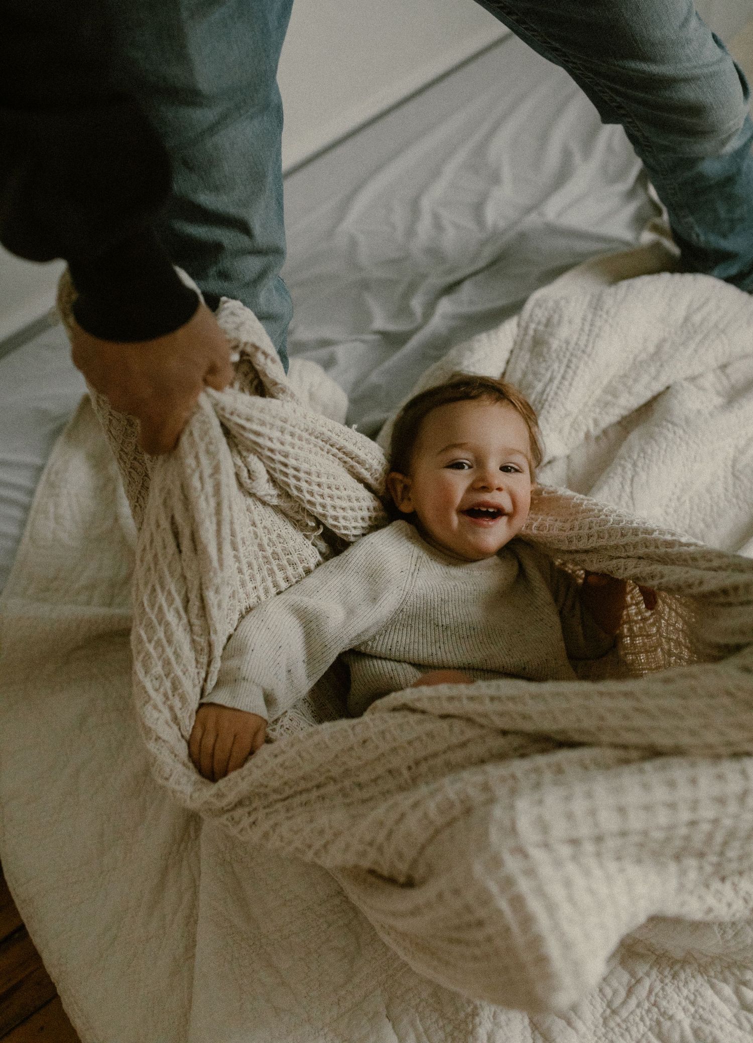 A laughing baby wrapped in knitted blankets on white bedding looks up with joy.