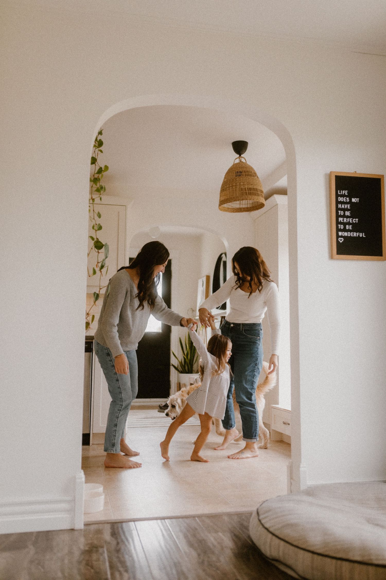 Queer family dancing together in a bright minimalist home interior with an arched doorway and woven pendant light.