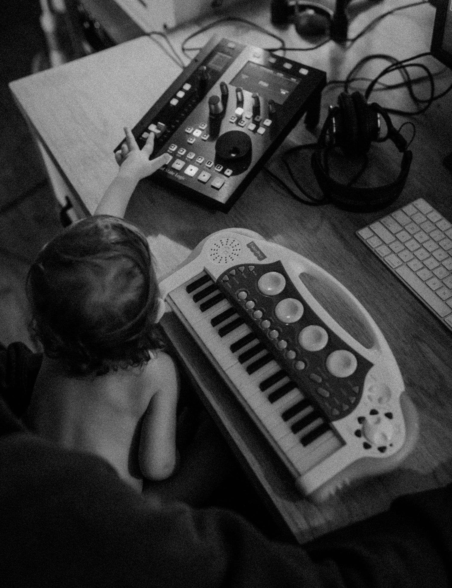 Black and white image showing a toddler sitting at a music production setup with a small keyboard synthesizer and audio interface.