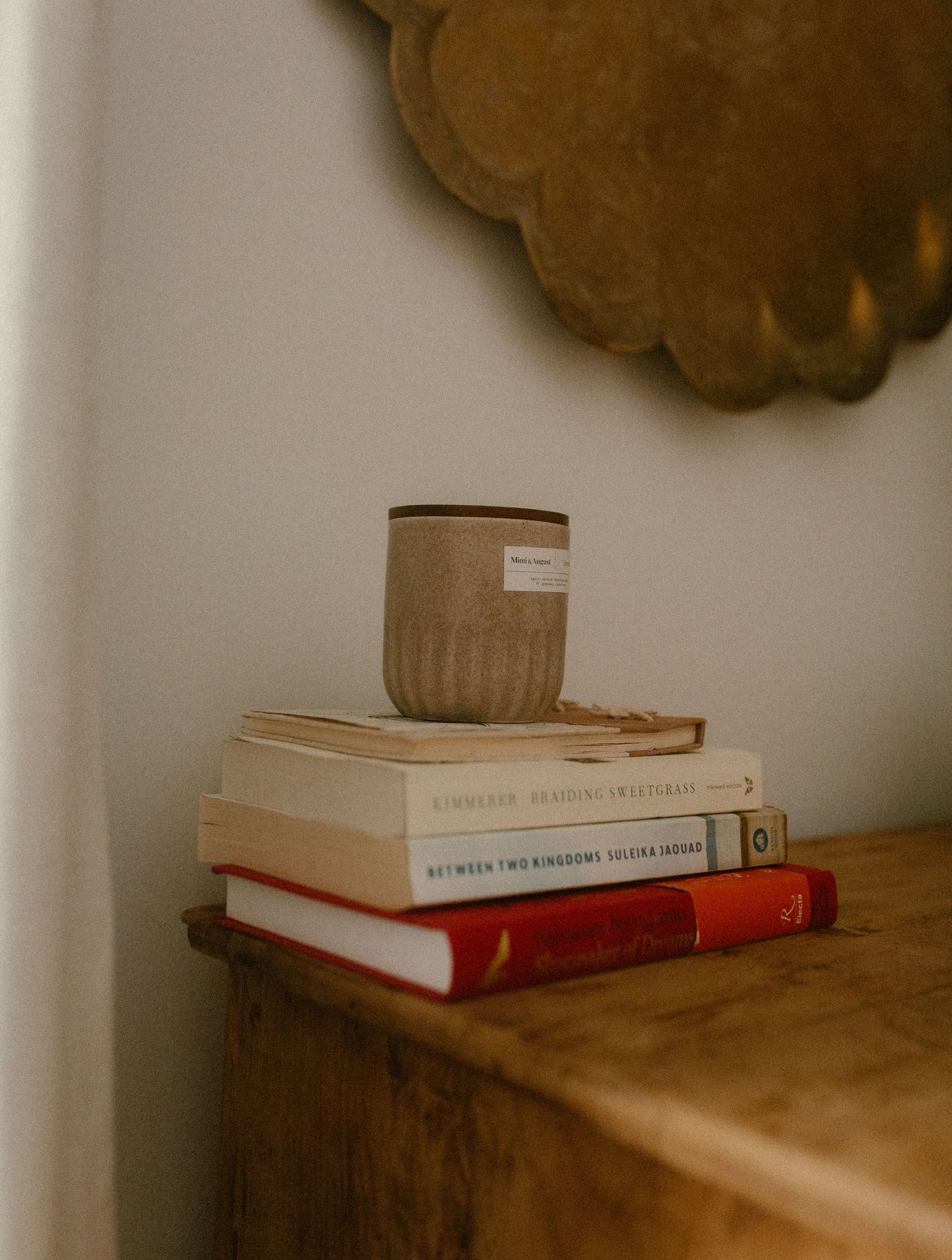 Stack of books with a ribbed ceramic mug on wooden surface beneath decorative wall art.