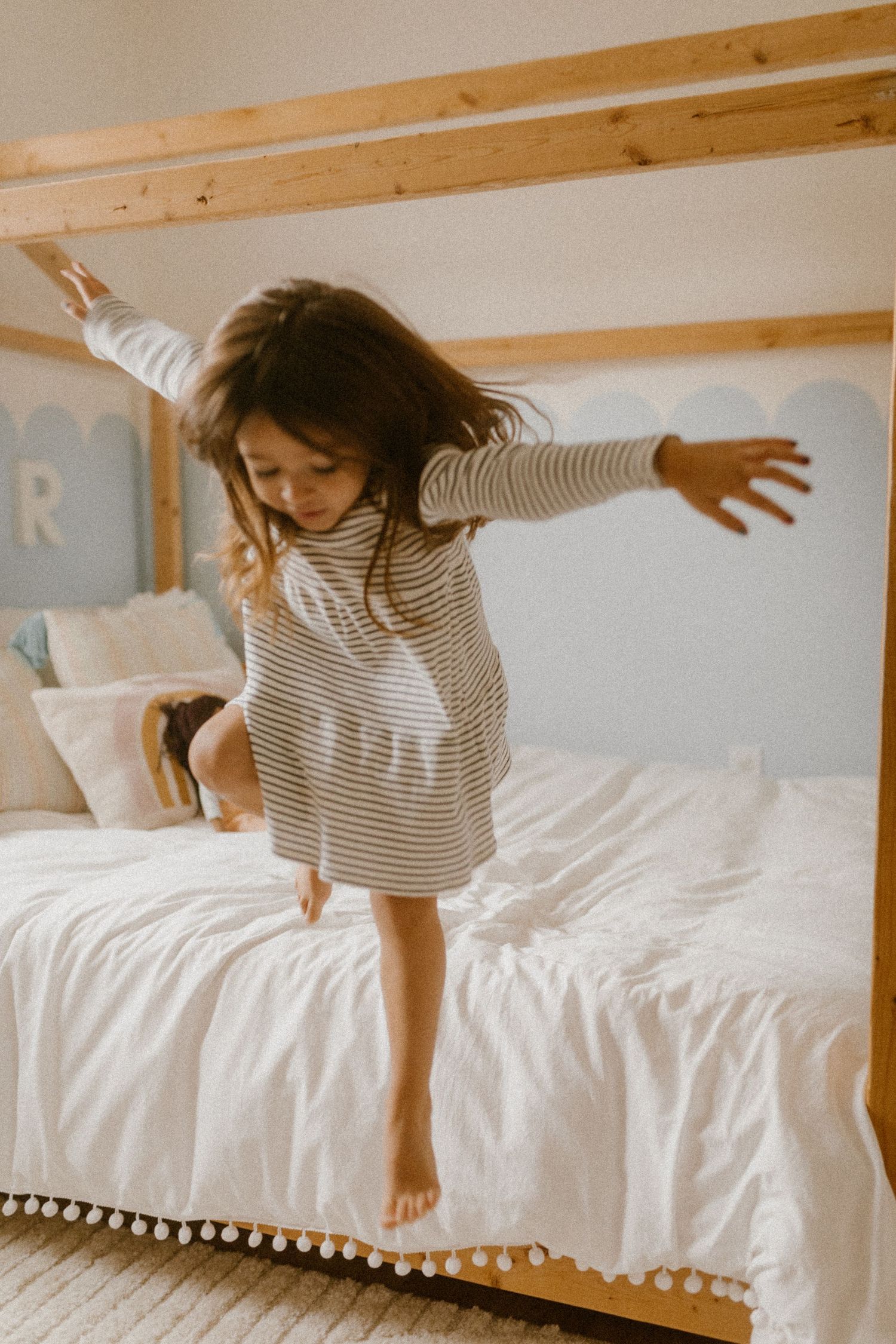 A child in a striped dress jumps playfully on a white bed with pom-pom trim beneath wooden beams.