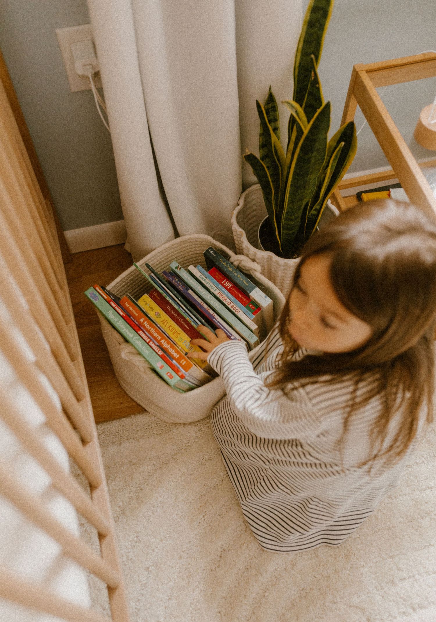 Small child in striped dress reaches into storage bin filled with children's books near a house plant.