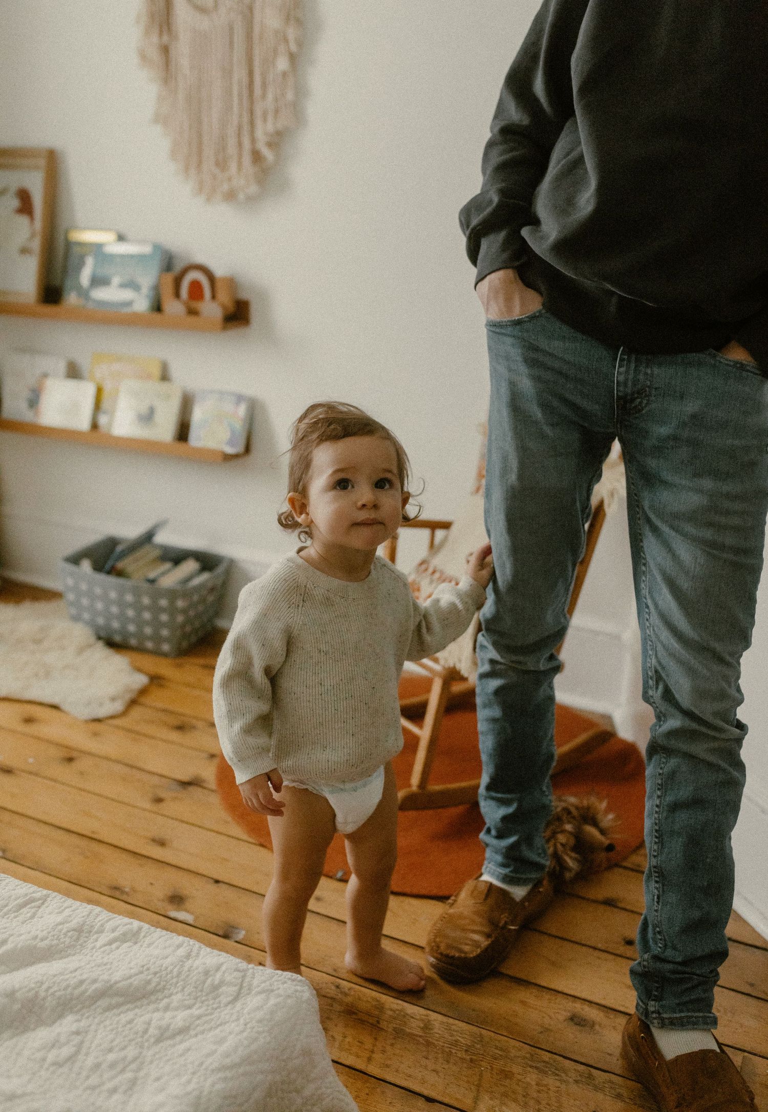 A toddler in a cream sweater stands beside an adult on wooden flooring in a cozy room.