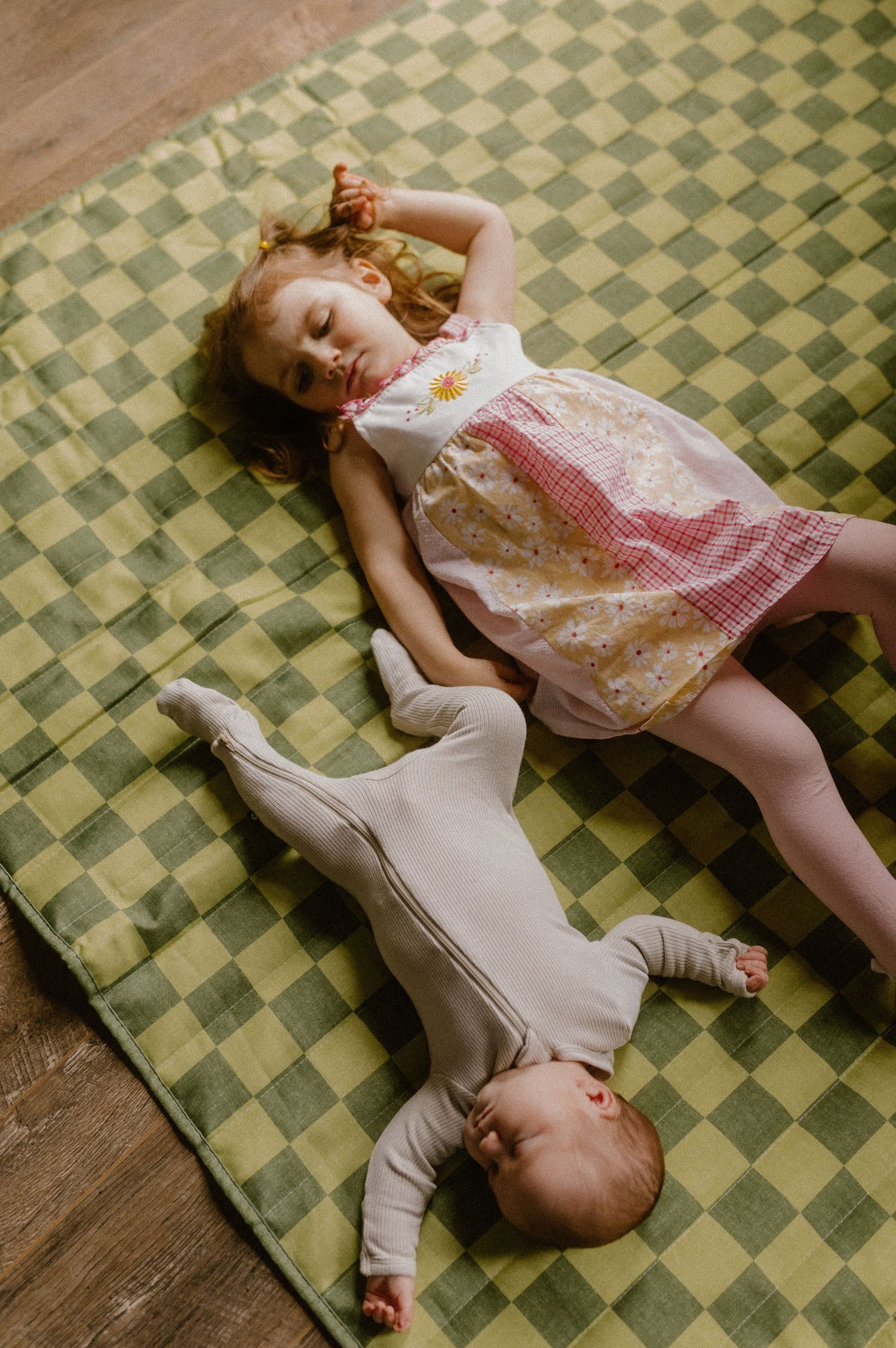 Sisters resting on a vintage checkered rug, one wearing a floral dress and the other in a light-colored onesie.