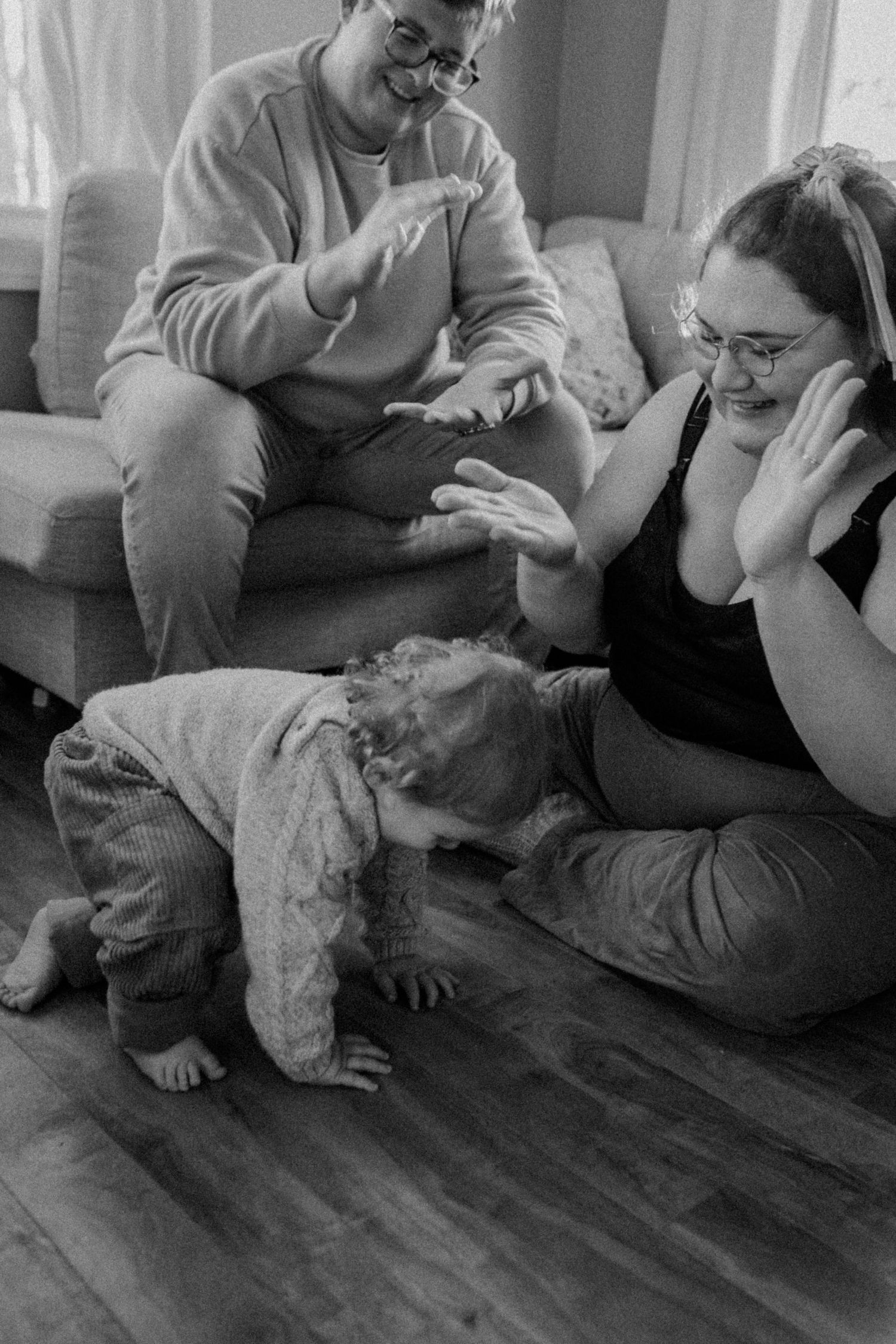 A candid black and white photo showing a toddler crawling on hardwood floors while fat parents share a laugh on the couch.