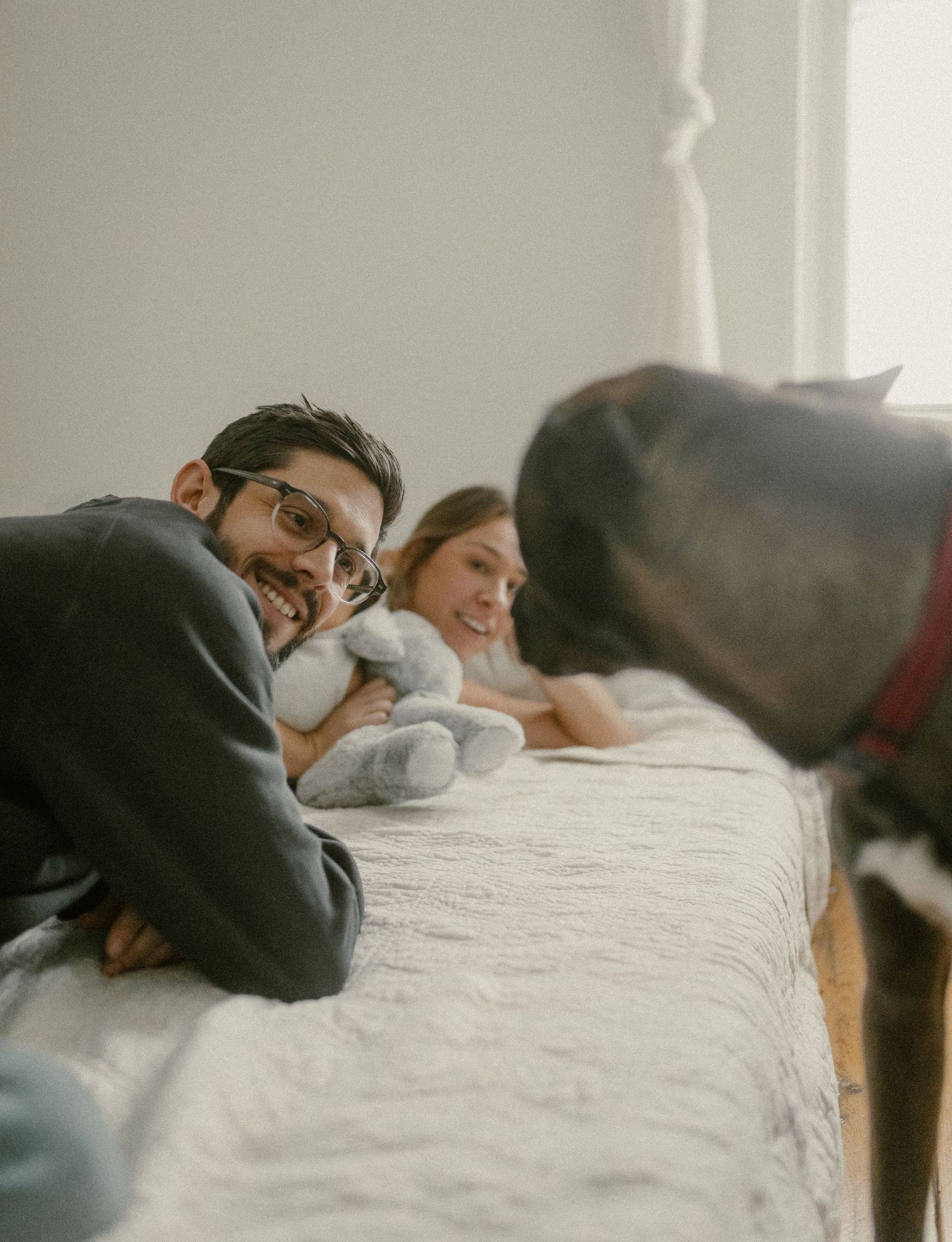 Family lounging on bed while black dog peeks into frame from the side.