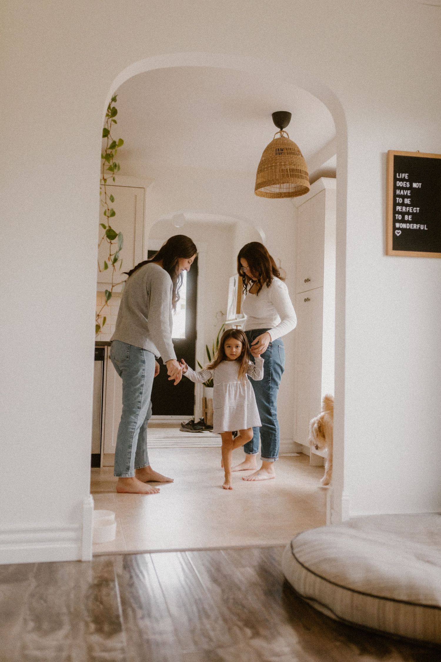 Intimate queer family dancing in bright hallway with arched doorways and woven light fixture.