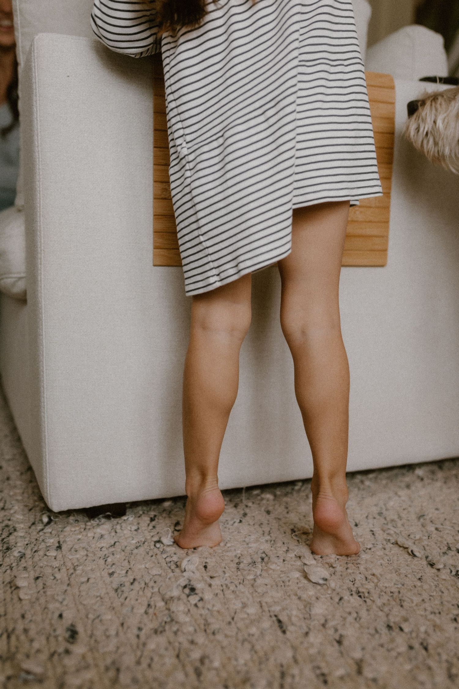 Child on her tippy toes in a striped dress against a light gray couch on a carpeted floor.