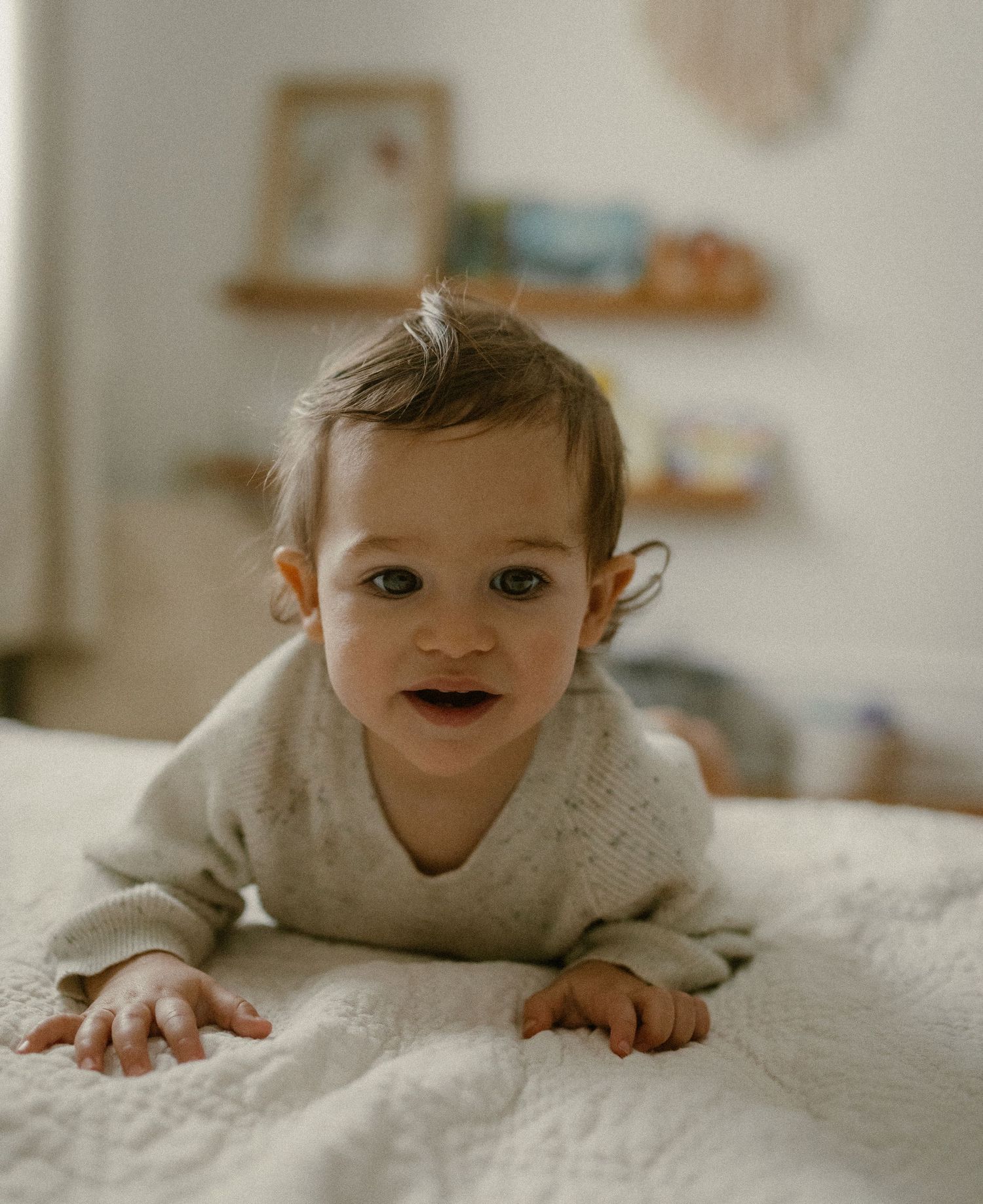 Close-up photo of a toddler on white bedding with floating shelves and wall decor in background.