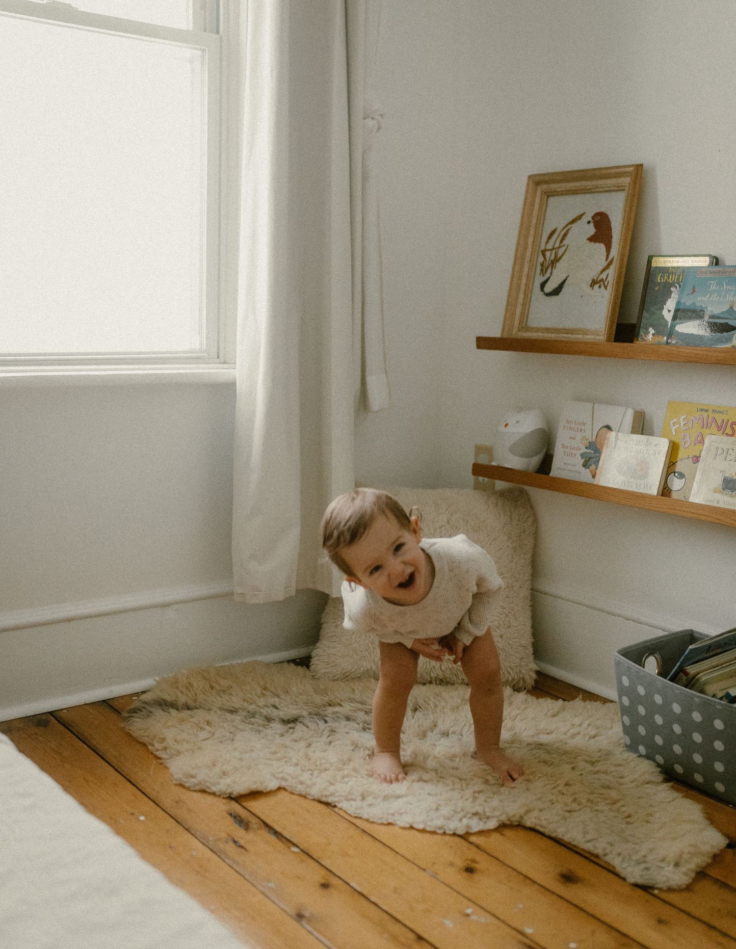 A toddler plays on a circular rug near a window with artwork displayed on floating shelves.
