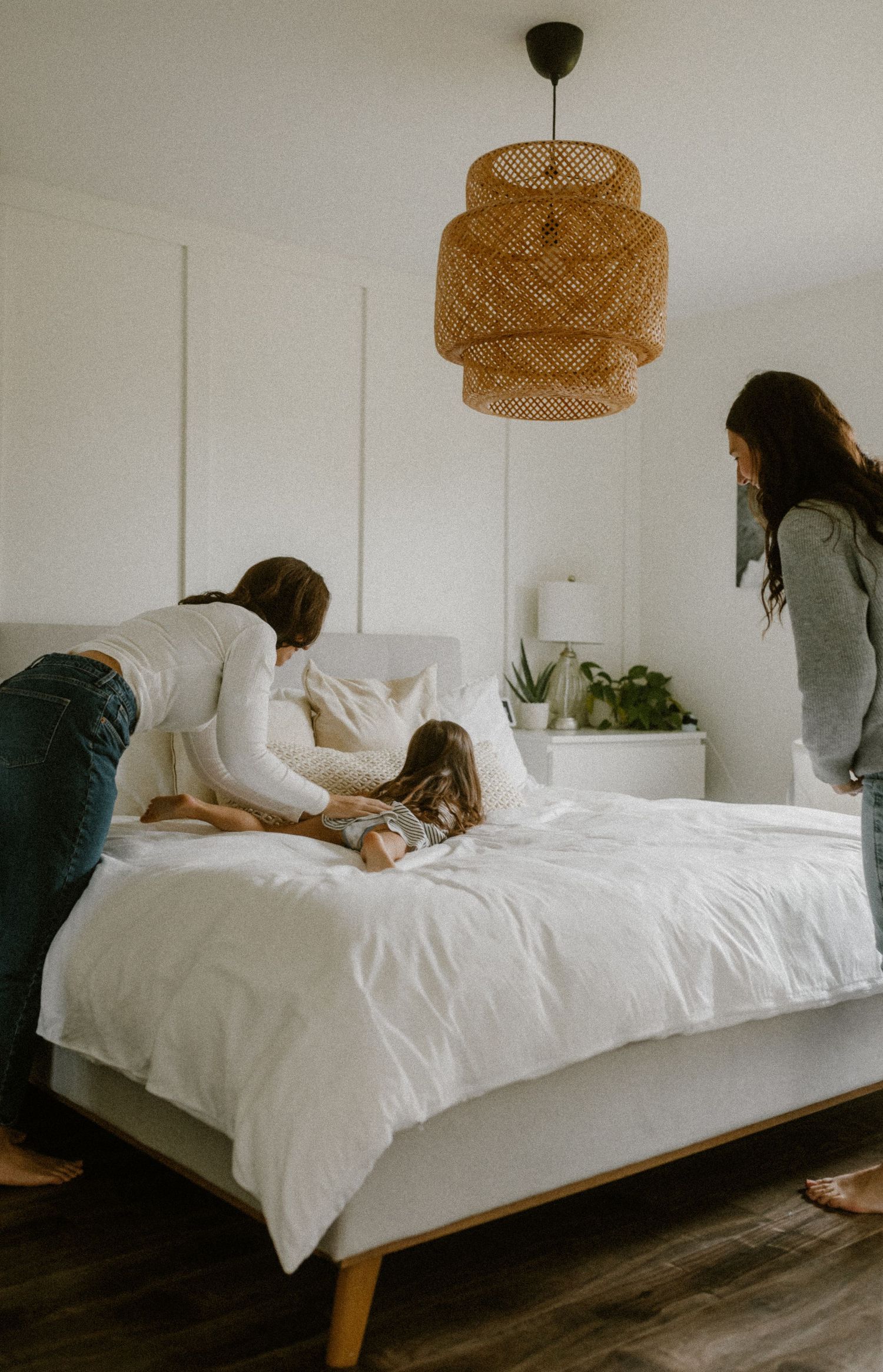 People playing on a bed with crisp white linens under a woven rattan pendant light in a minimalist bedroom.