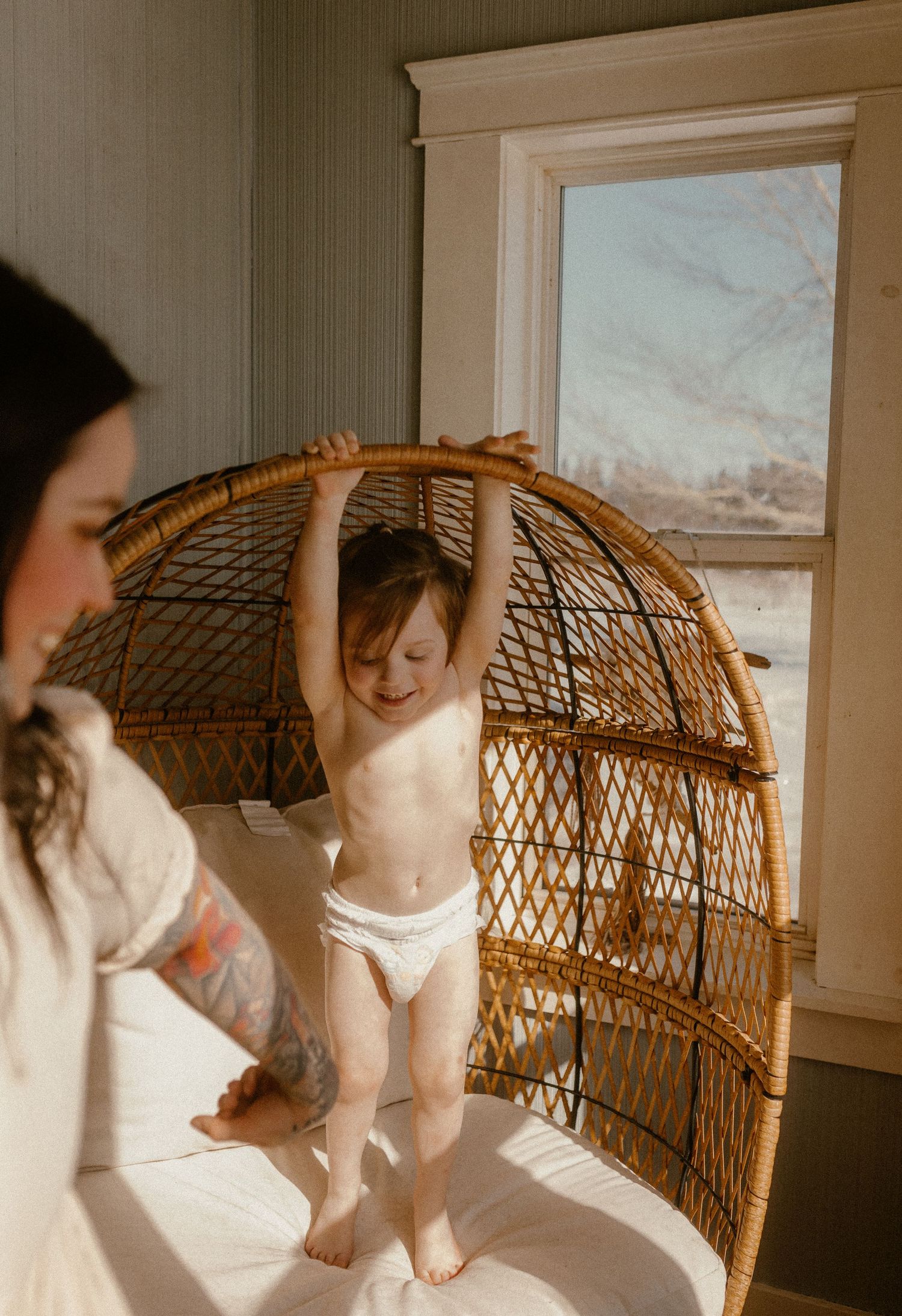 A toddler in a diaper hangs playfully from a vintage wicker peacock chair near a window bathed in natural light.