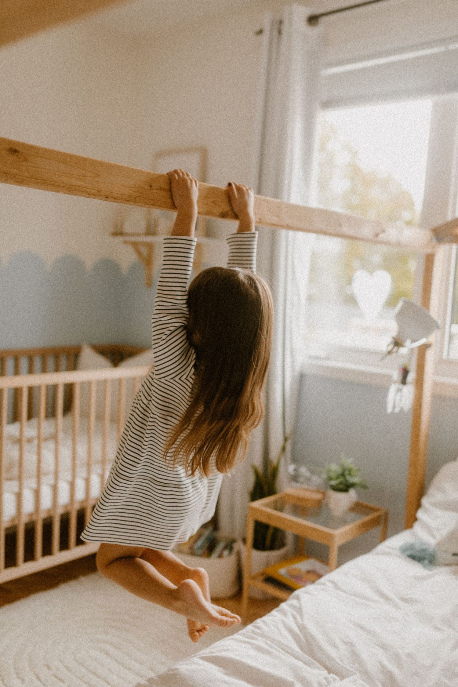 Child in striped shirt stretches towards wooden beam in cozy bedroom with crib and natural light through window.