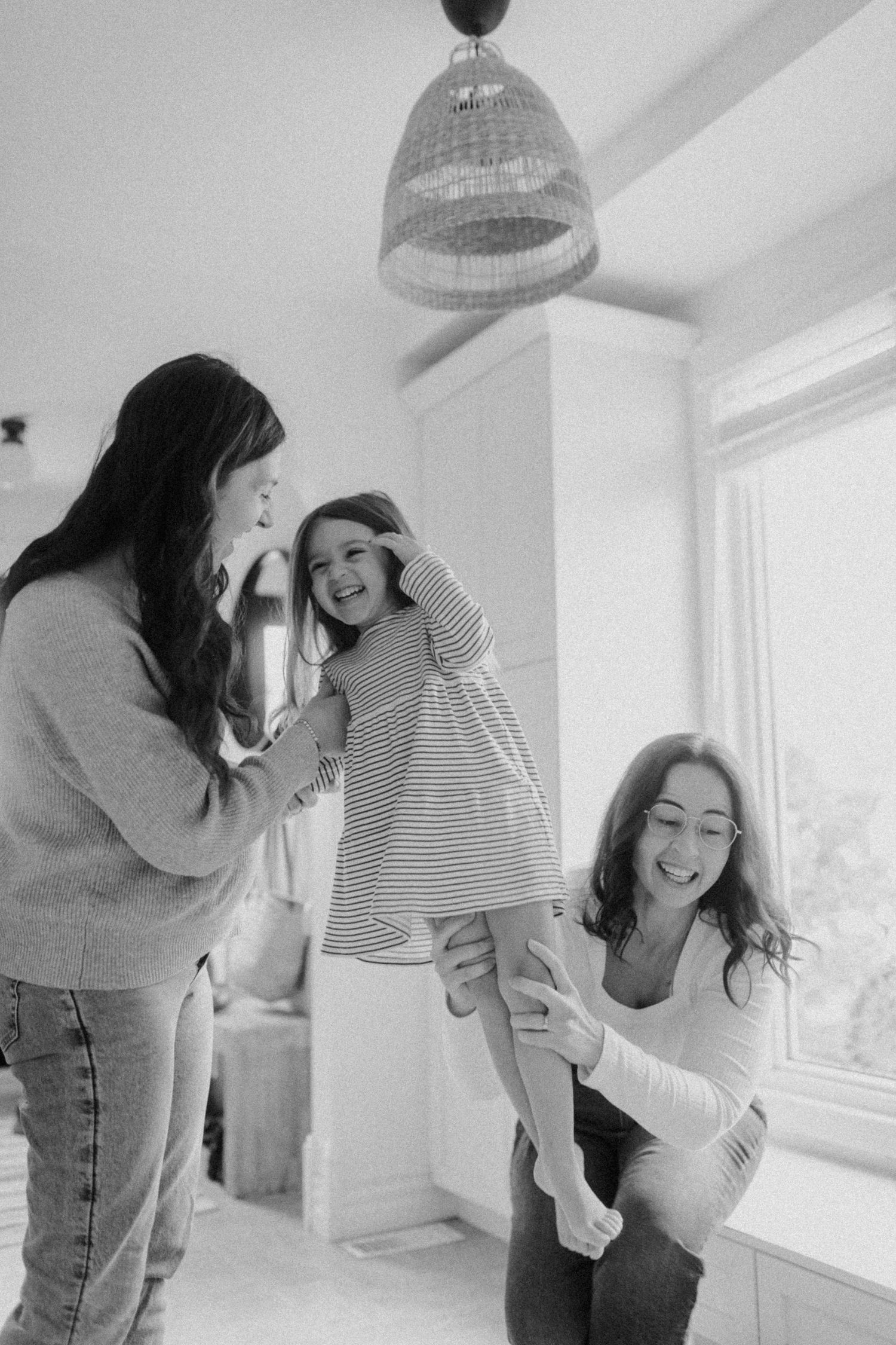 A black and white candid photo showing joyful playtime as they lift and swing a small child in a striped dress.