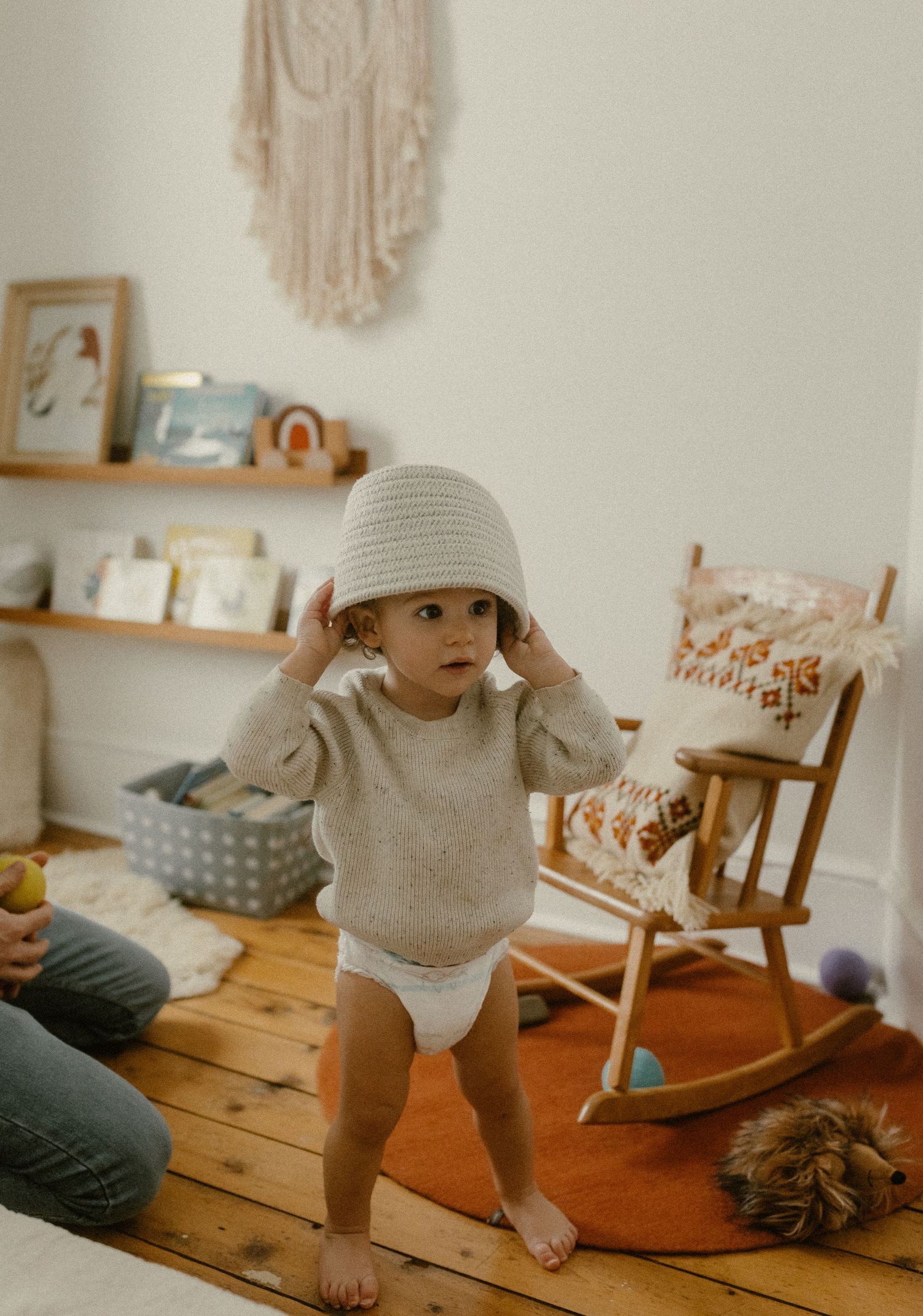 A silly toddler wearing a basket as a hat and sweater stands on wooden floors in a minimalist nursery.