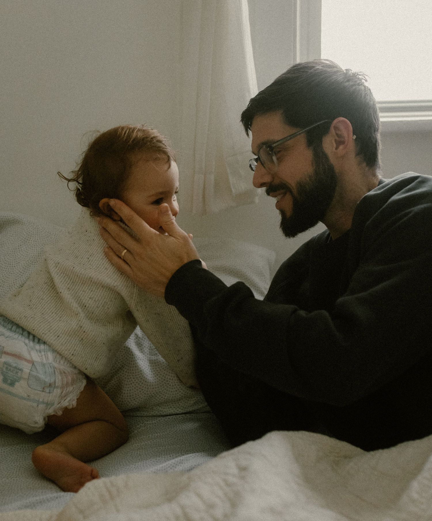 An intimate moment captured in soft lighting as father and daughter share a tender interaction on white bedding.