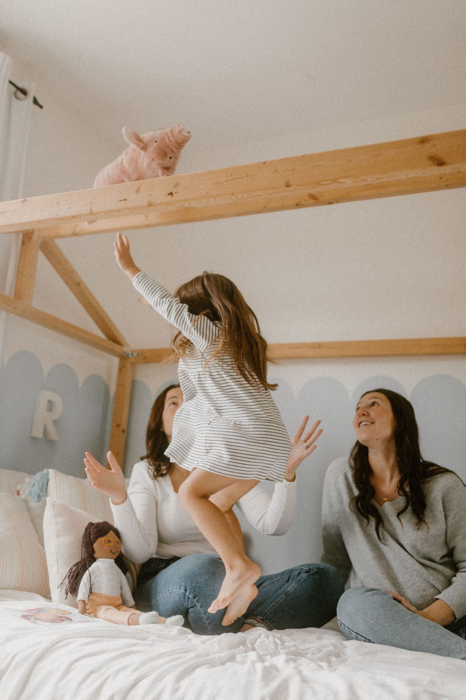 Little girl jumps on a bed with stuffed animals and wooden beam details as her two moms watch.