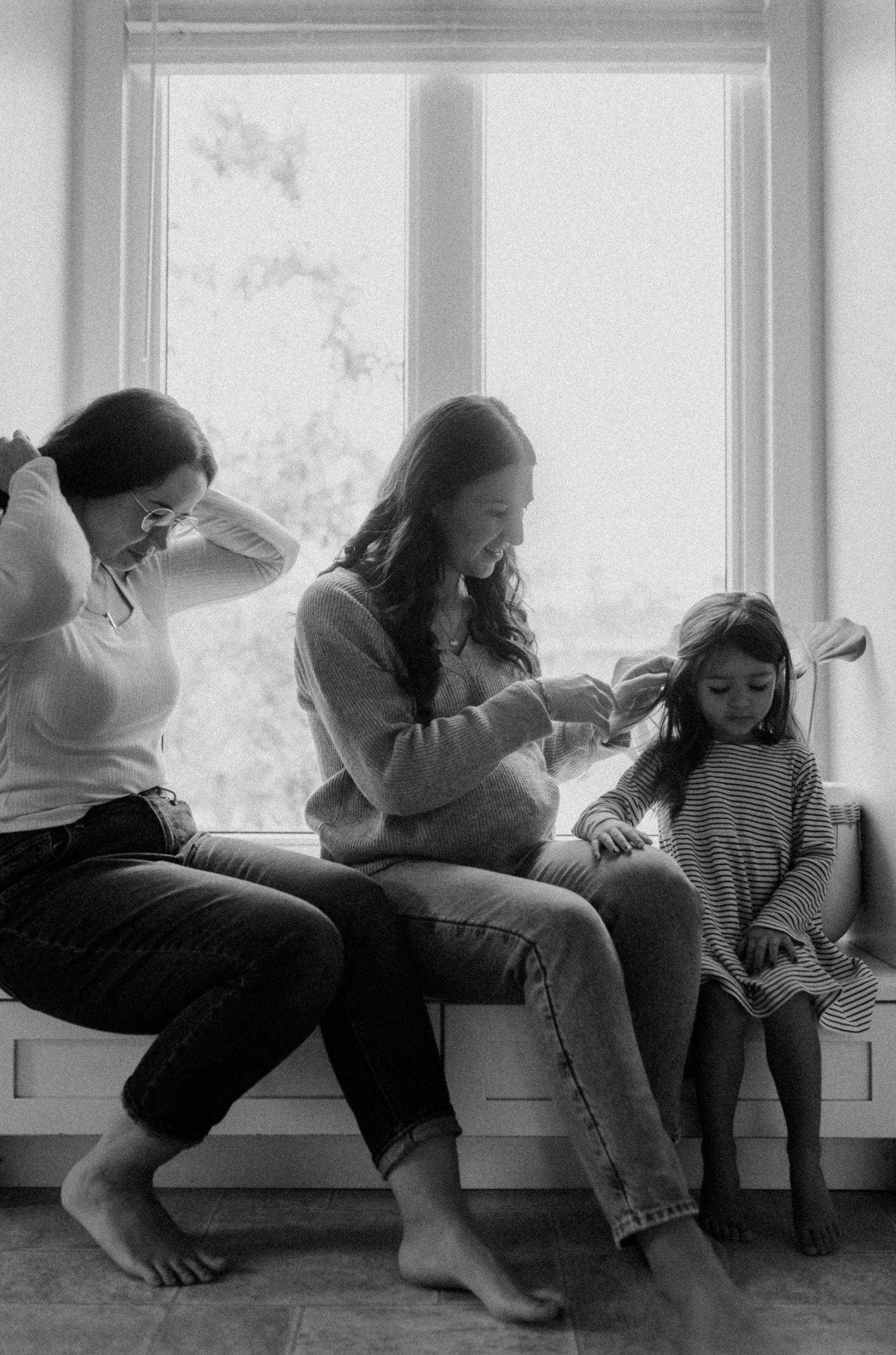 A queer family sit casually by a bright window in a candid black and white photograph showing a tender bonding moment.