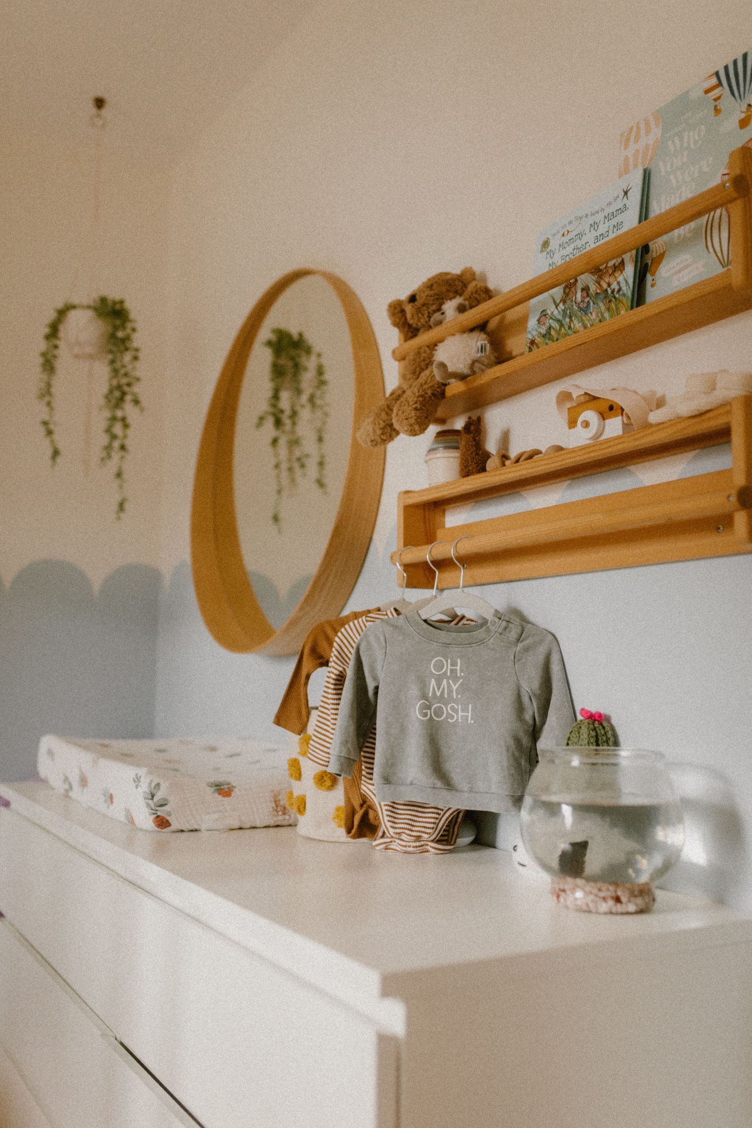 A cozy nursery dresser with round mirror, hanging plants, and adorable stuffed animals on display.