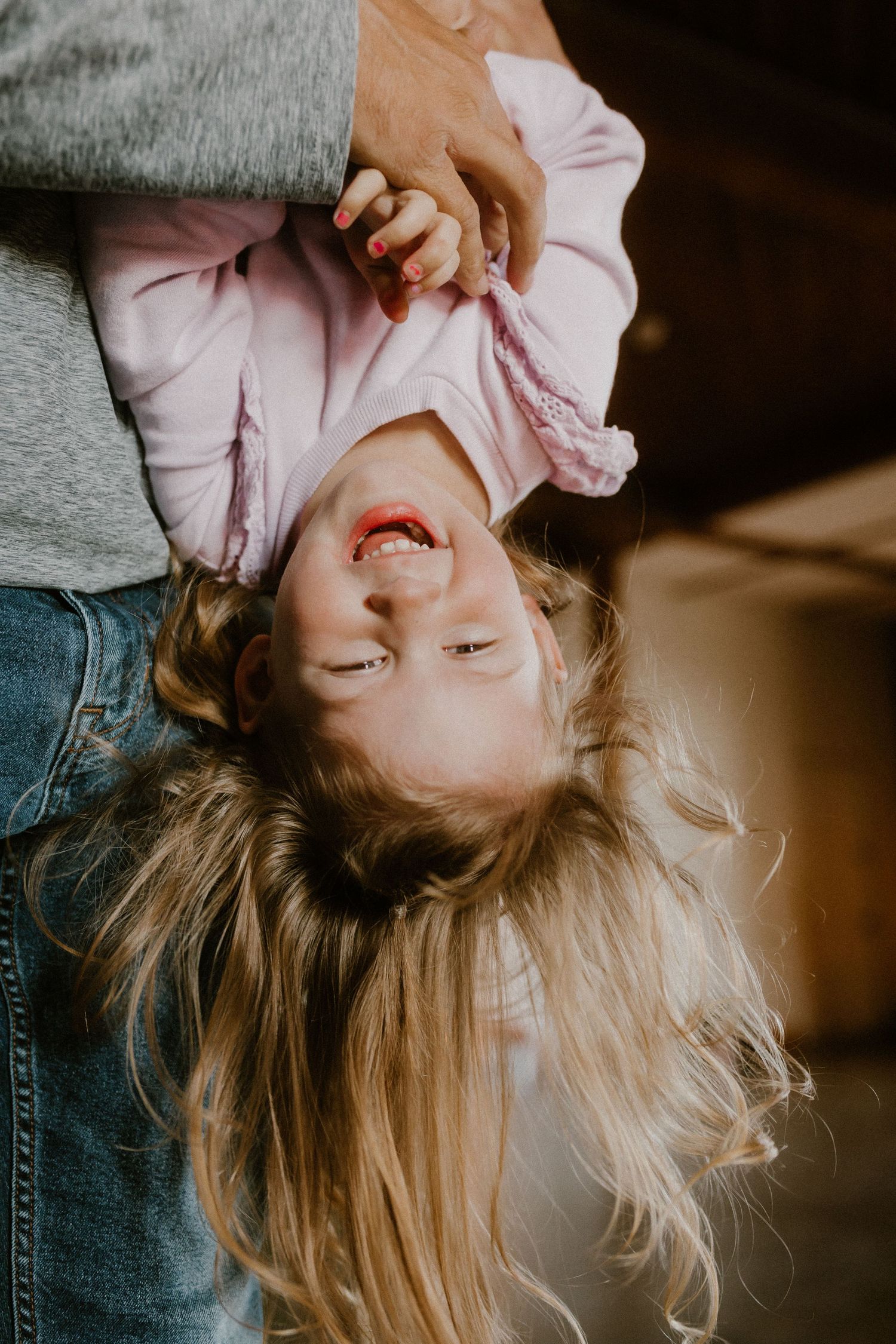 A young child with flowing blonde hair dangles upside down while being held, laughing joyfully.