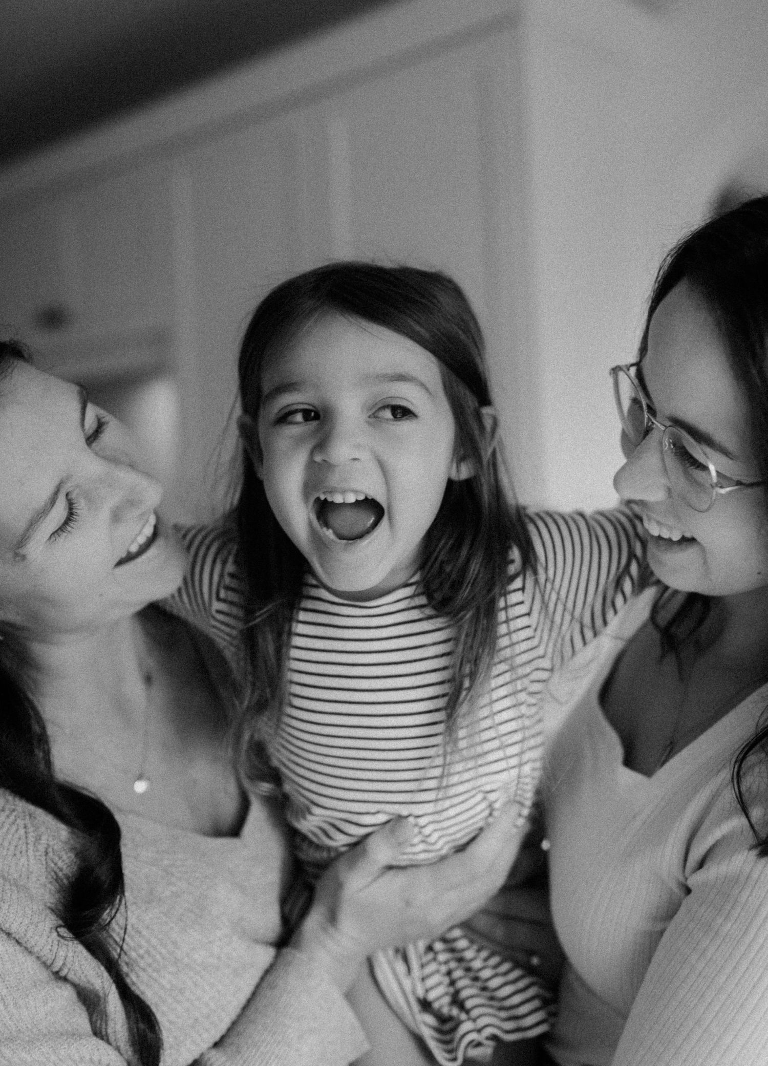 A joyful black and white candid queer family moment showing spontaneous laughter and embraces in an intimate indoor setting.