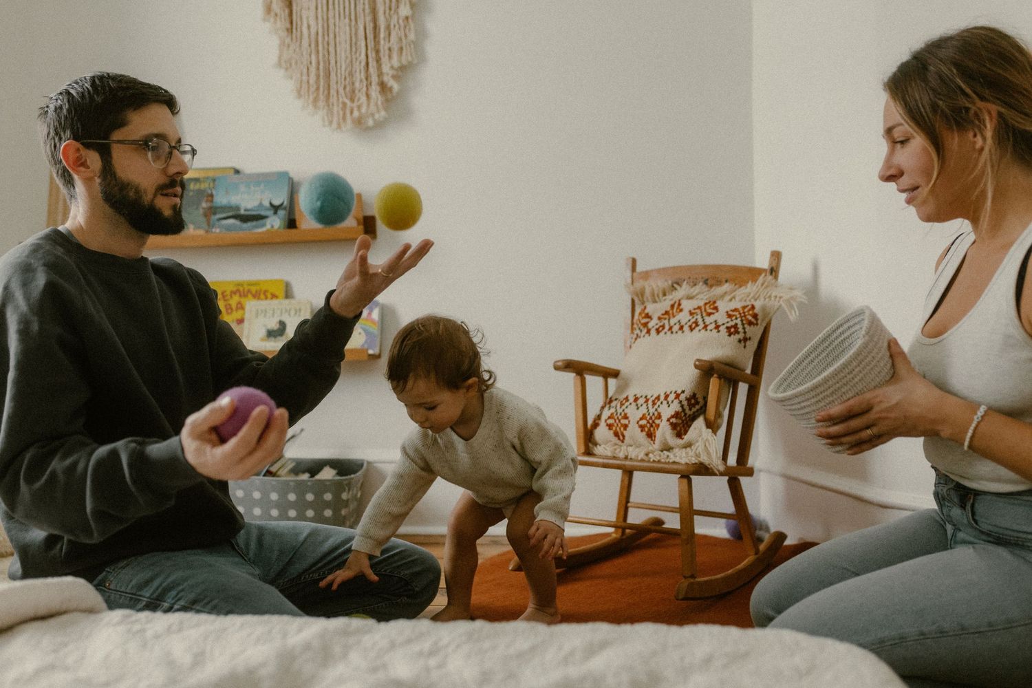 Family sitting on bed playing with colorful balls in cozy bedroom with macrame wall hanging.