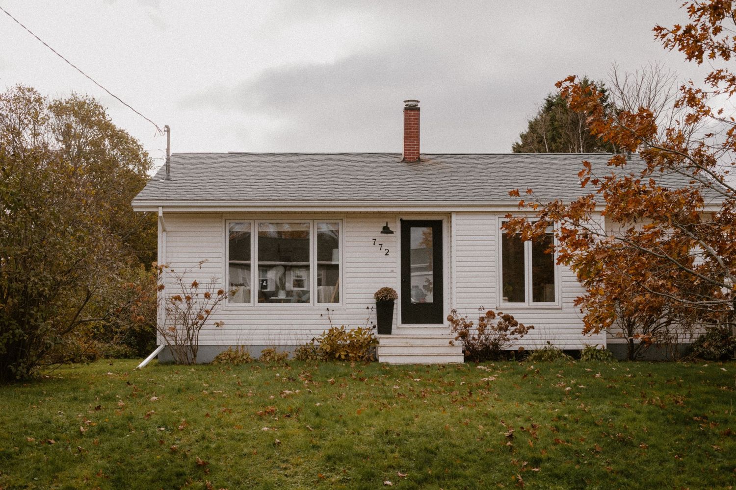 A charming white cottage house with grey roof and autumn trees in the background in Summerside, PEI.