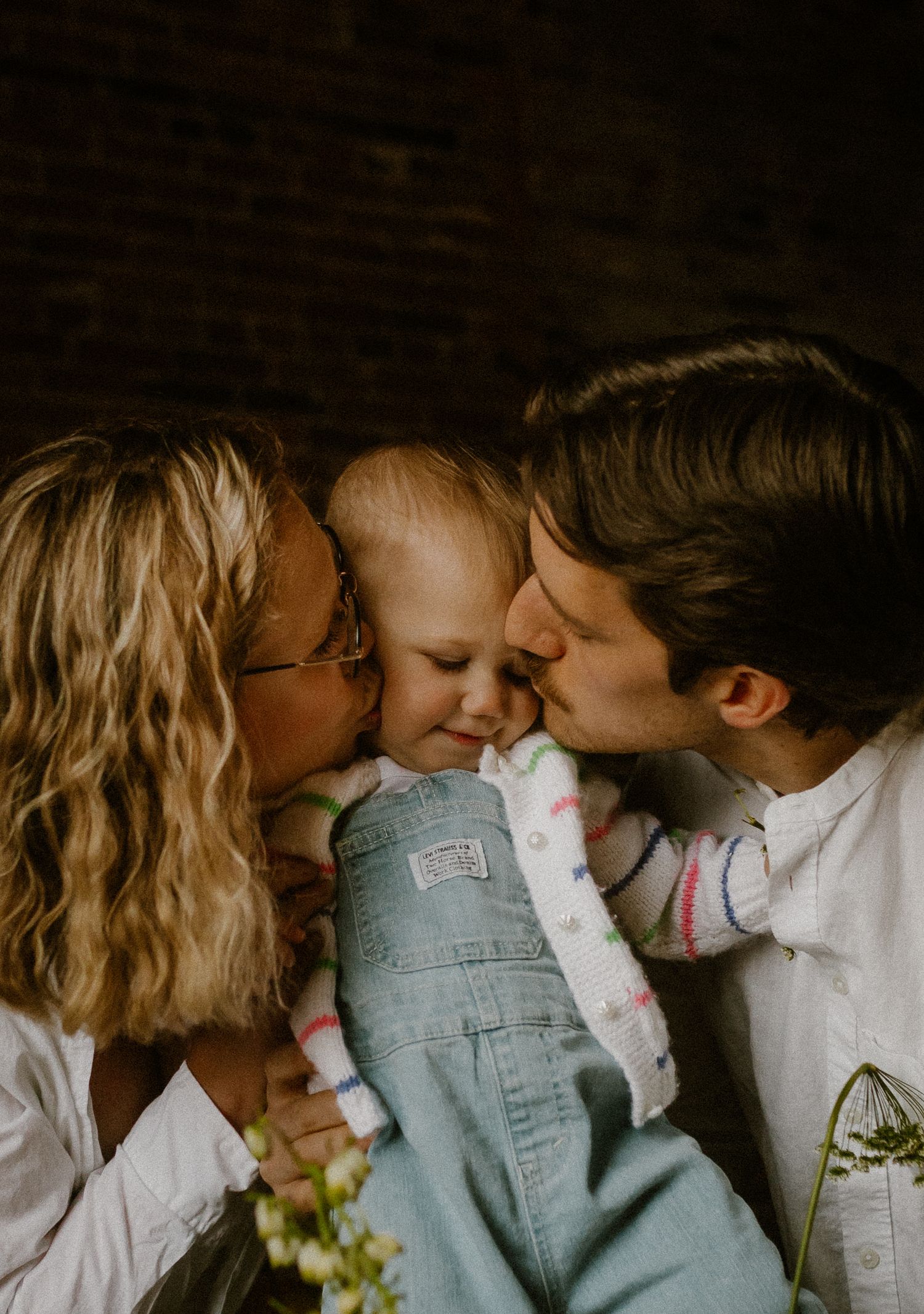 Sweet family moment showing parents kissing their baby in overalls while sharing an intimate embrace together.