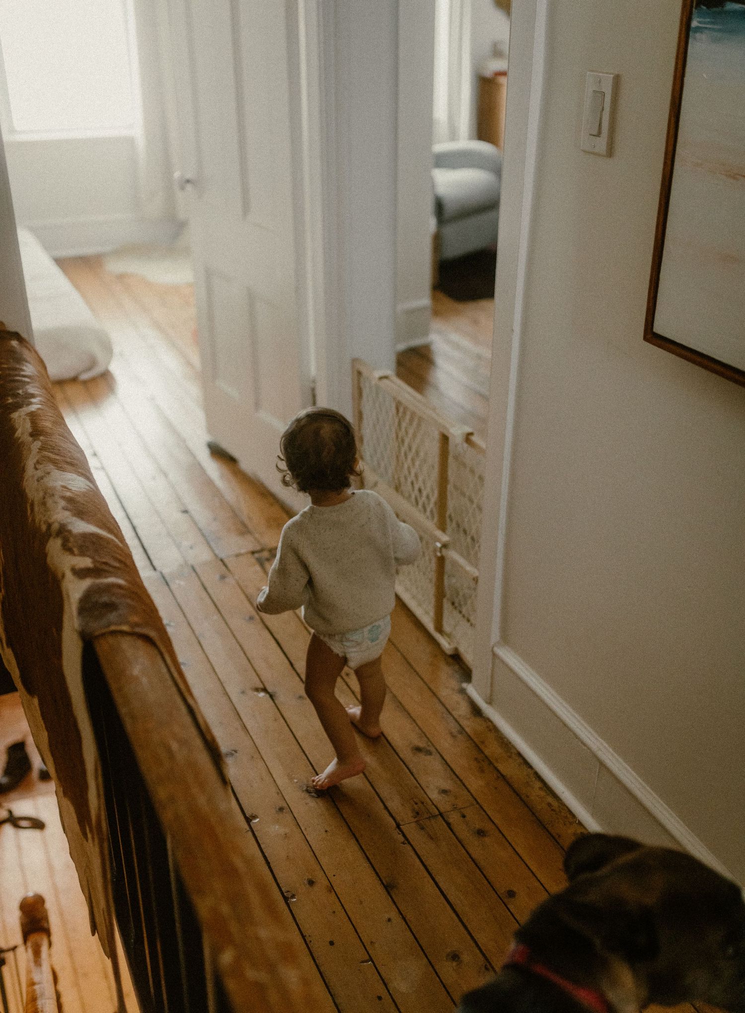 Small child walking along a rustic wooden hallway with white walls and visible baseboards.