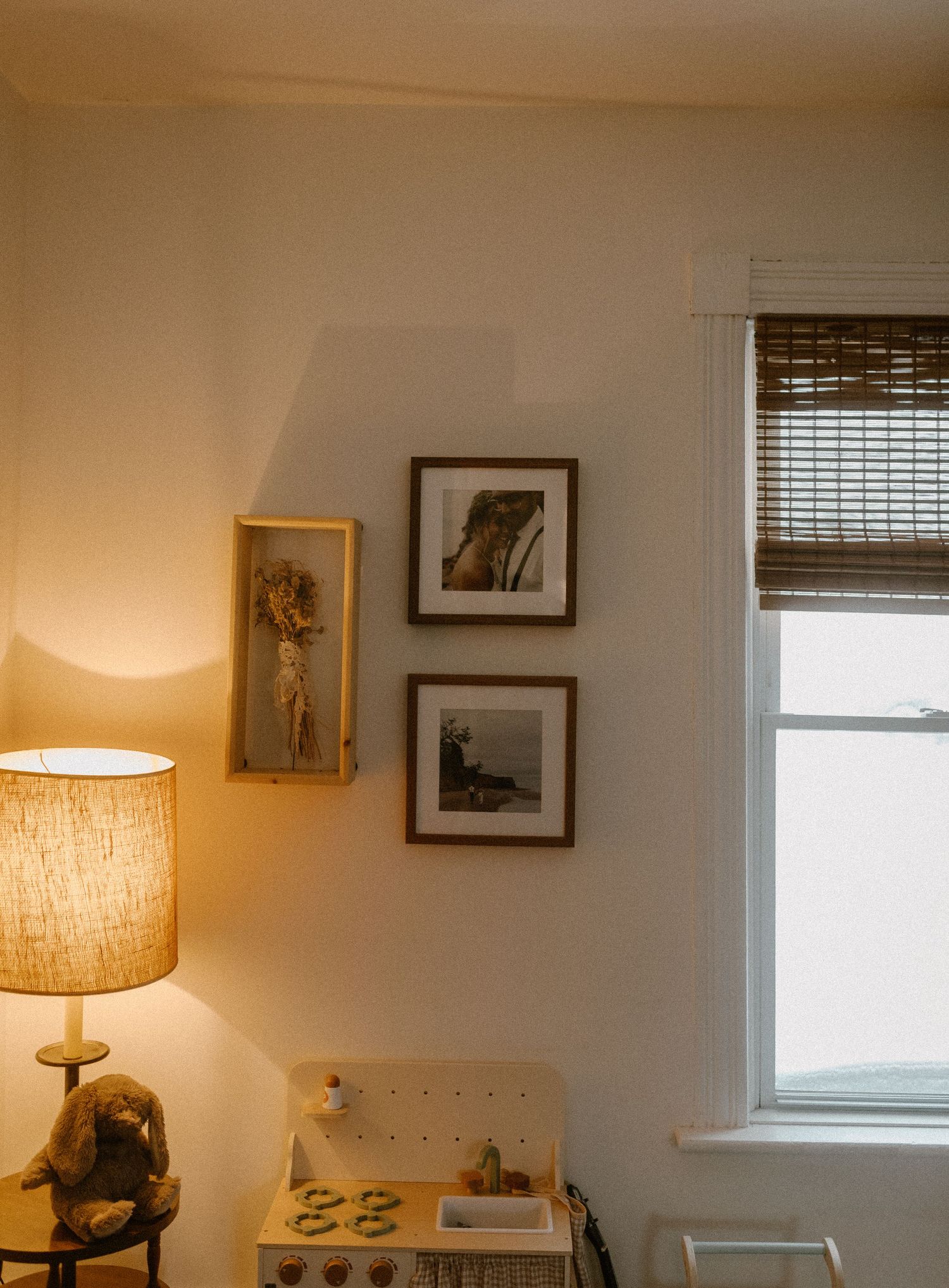 Cozy corner of a room with framed artwork, a window, and a lit table lamp creating warm ambient lighting.