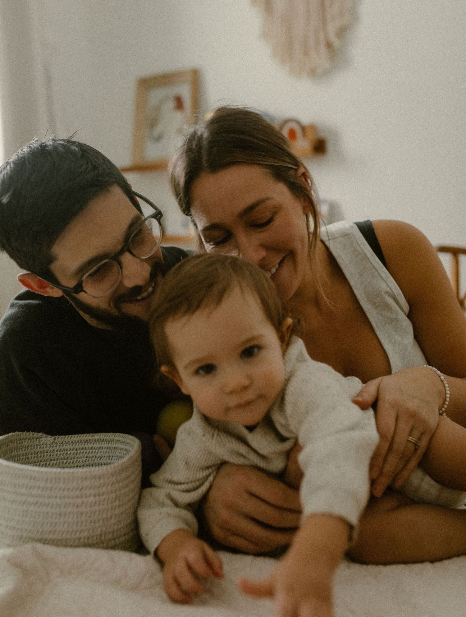 A family shares an intimate moment together while relaxing on a bed with neutral decor and wall hangings in the background.
