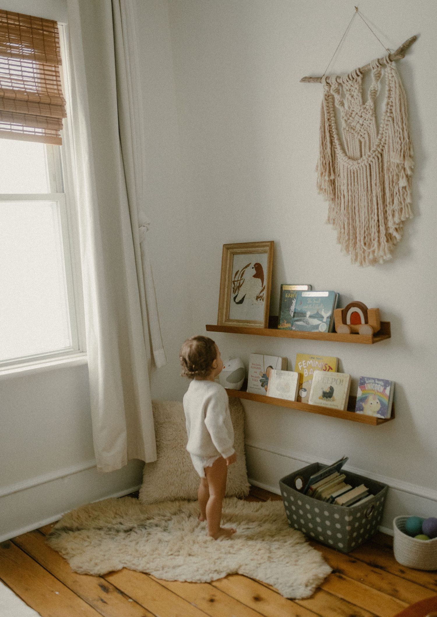 Small child stands on a white rug looking at artwork and macrame wall hanging in a sunlit room.