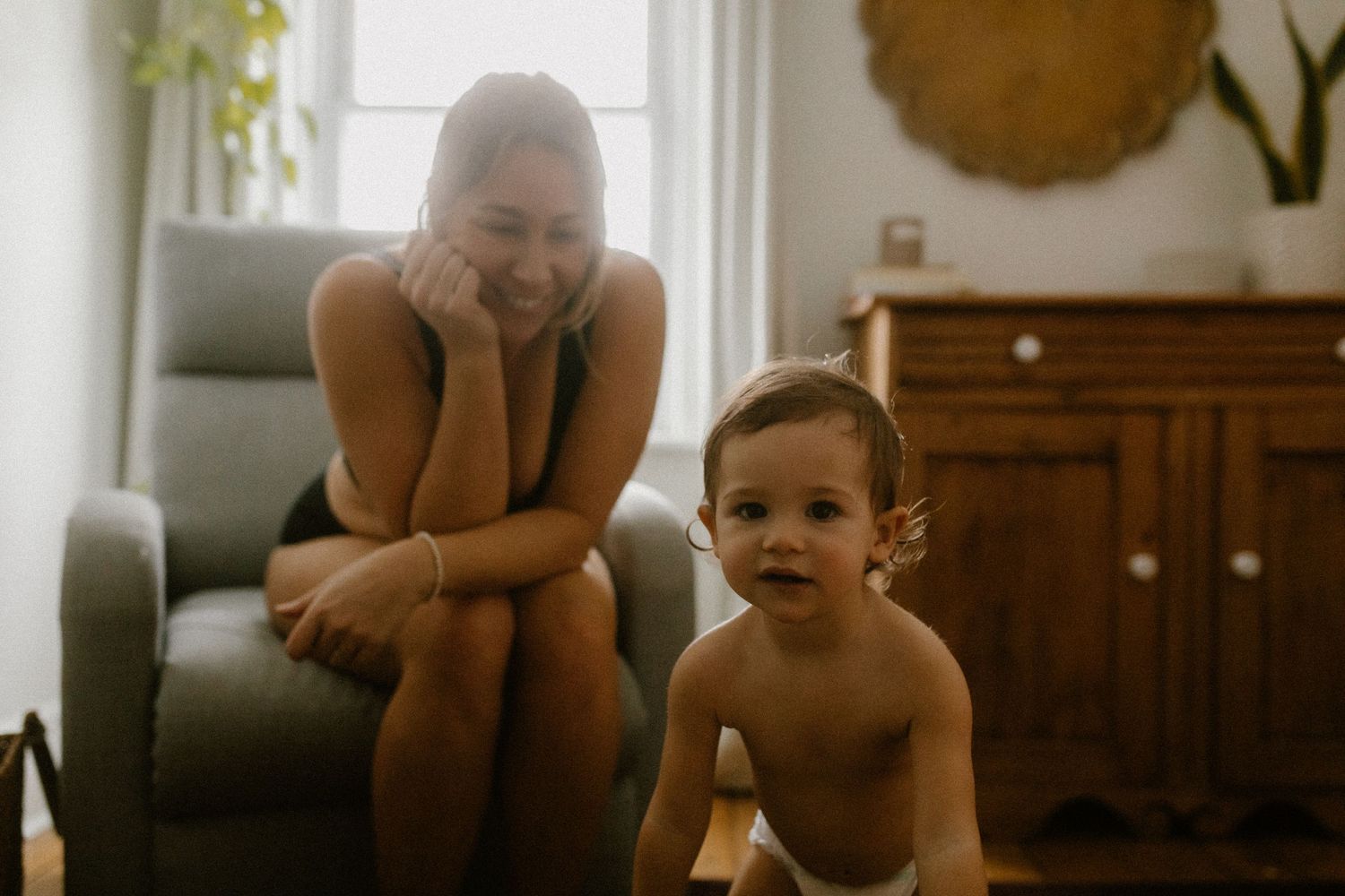 A joyful indoor scene shows a toddler sitting on the floor beside a grey armchair in a cozy living room.