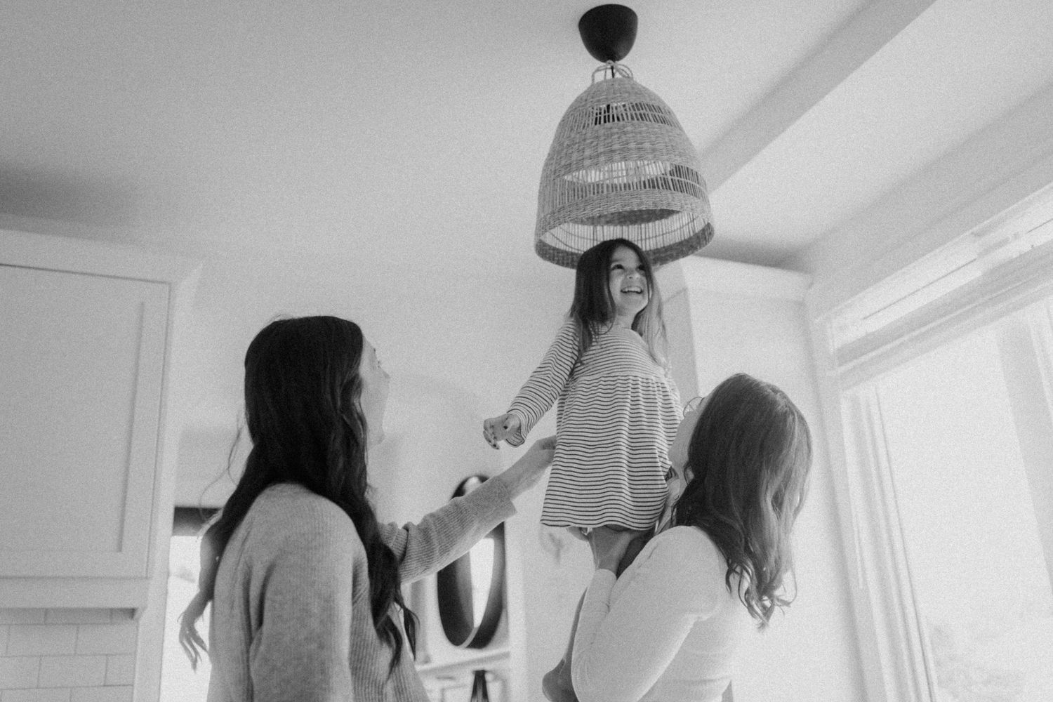 Black and white photo showing playful family interaction under a hanging pendant lamp in a bright room.