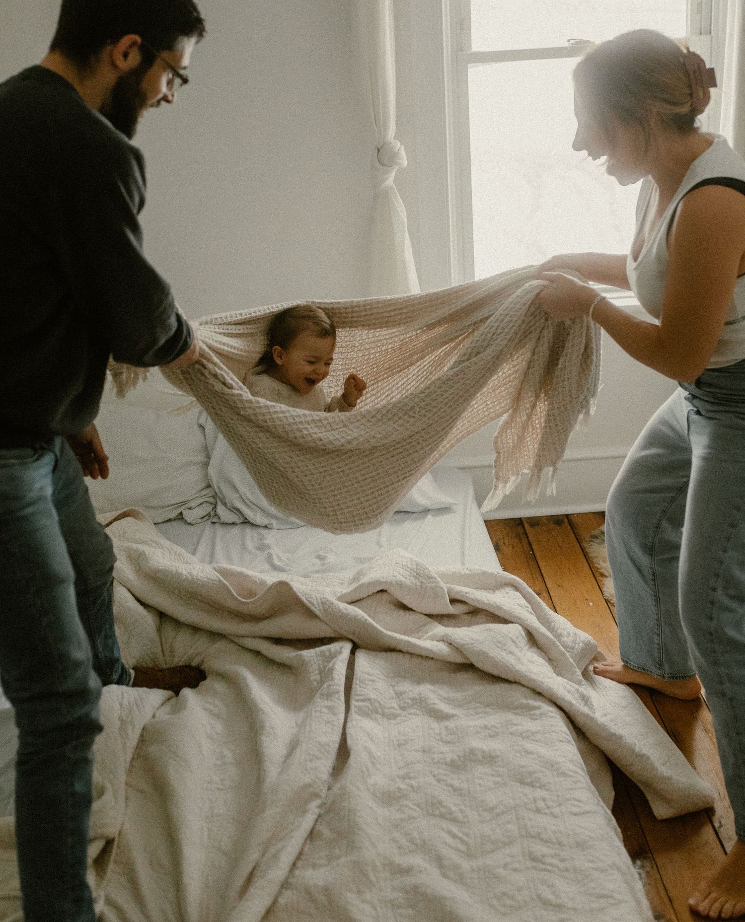 A couple gently swings their baby in a makeshift blanket hammock in a bright room.