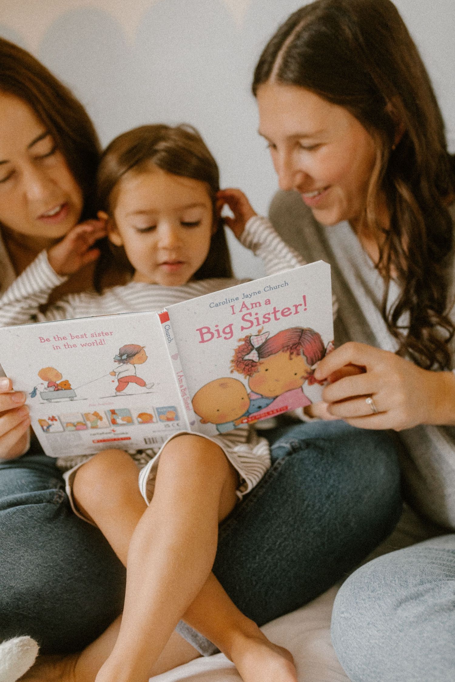Queer family reading 'I'm a Big Sister' book together on a comfortable couch.