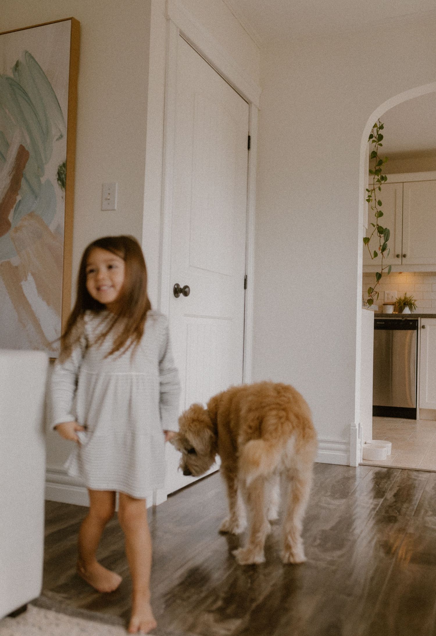 A child in a white dress walks barefoot with a golden doodle dog on hardwood floors.