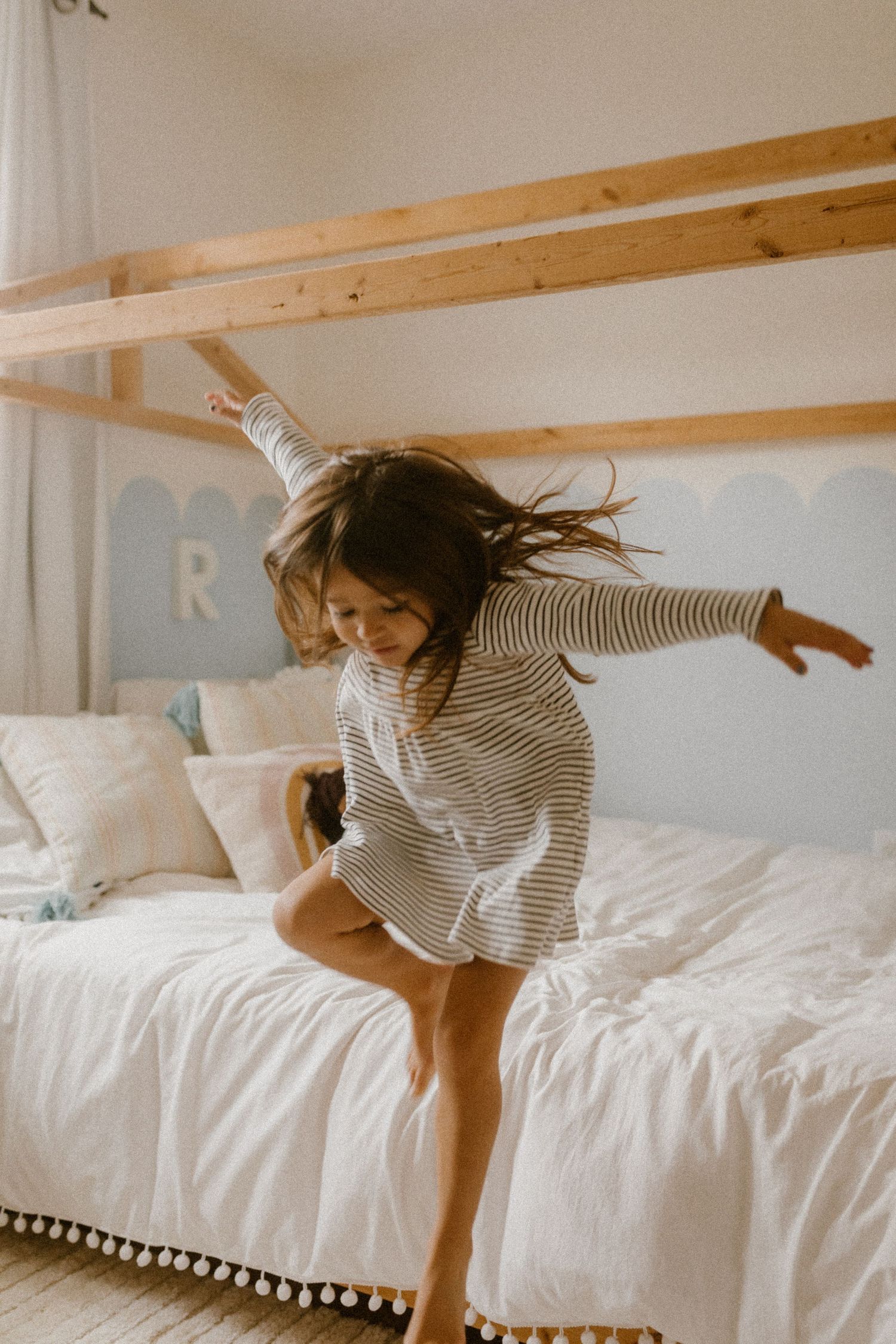 Young girl in striped dress jumps from white bed beneath wooden beams.